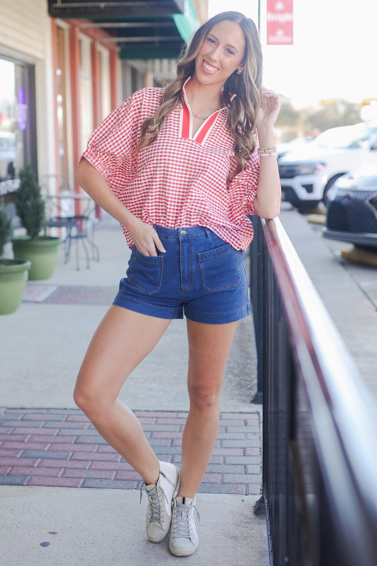 Woman wearing a red and white checkered top and blue shorts standing on a sidewalk.