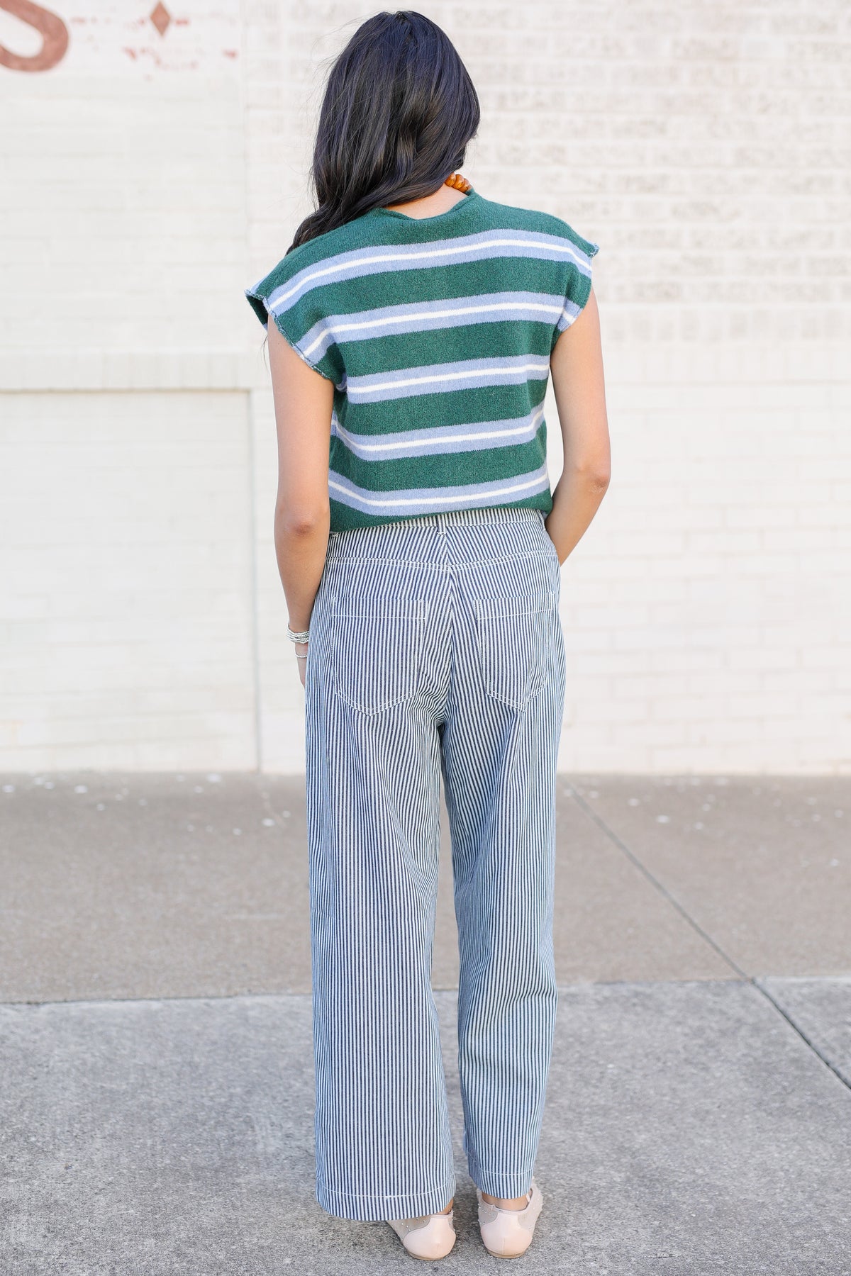 A woman is shown from behind wearing a green and white striped short-sleeve top and light blue pinstriped wide-leg pants, standing outdoors against a light brick wall.