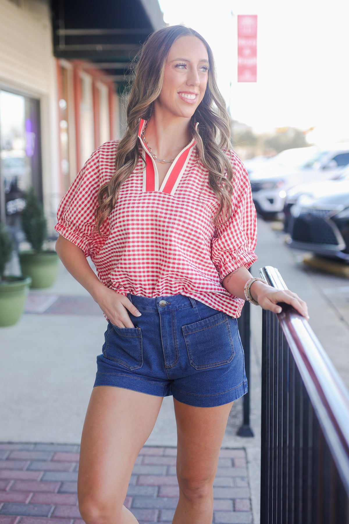 Woman wearing a red checkered shirt and blue shorts standing on a sidewalk.
