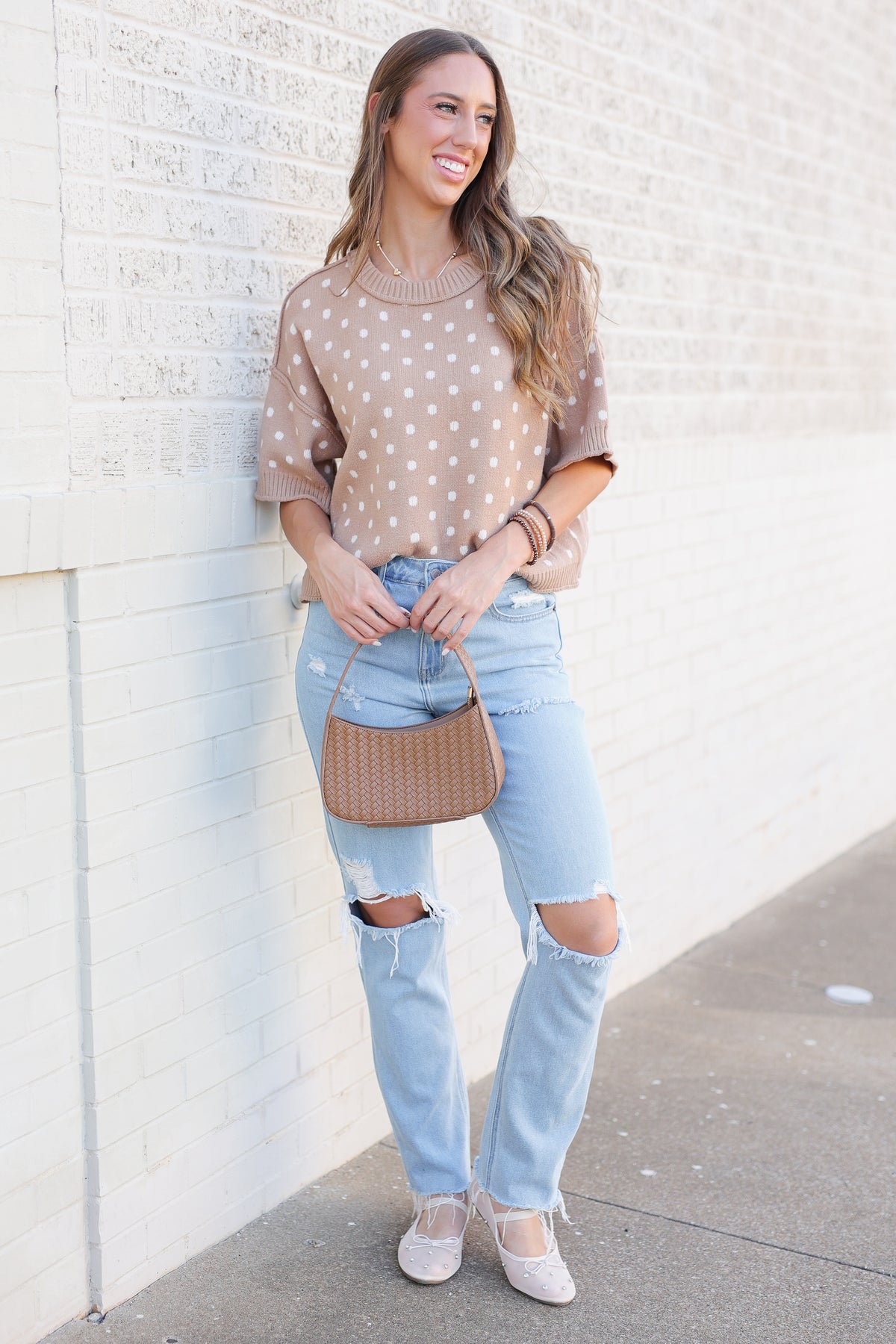 Blonde model standing outside near a brick wall wearing a camel colored sweater top with off white dots, blue jeans and flats.