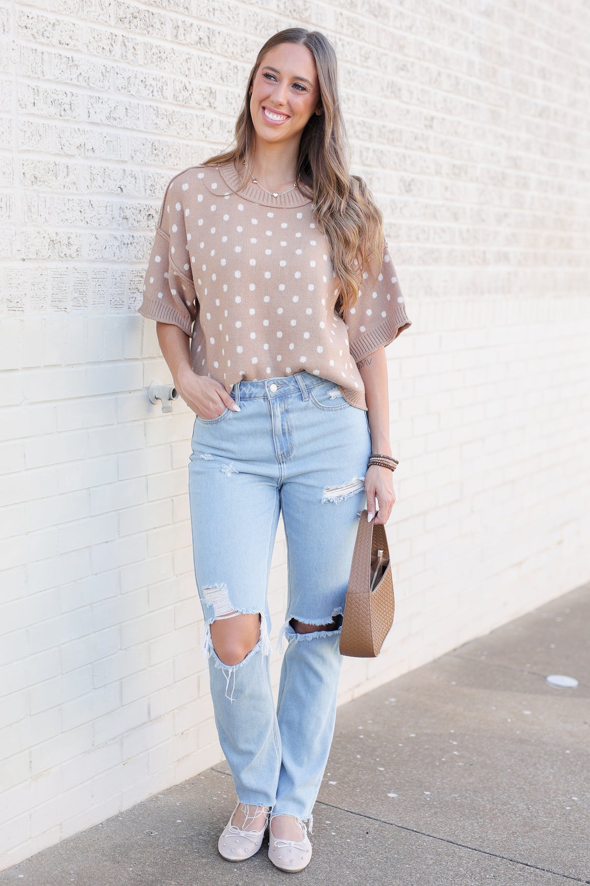 A woman stands against a white brick wall wearing a beige short-sleeve knit top with white polka dots, light-wash distressed jeans, and beige flats, while holding a brown handbag.
