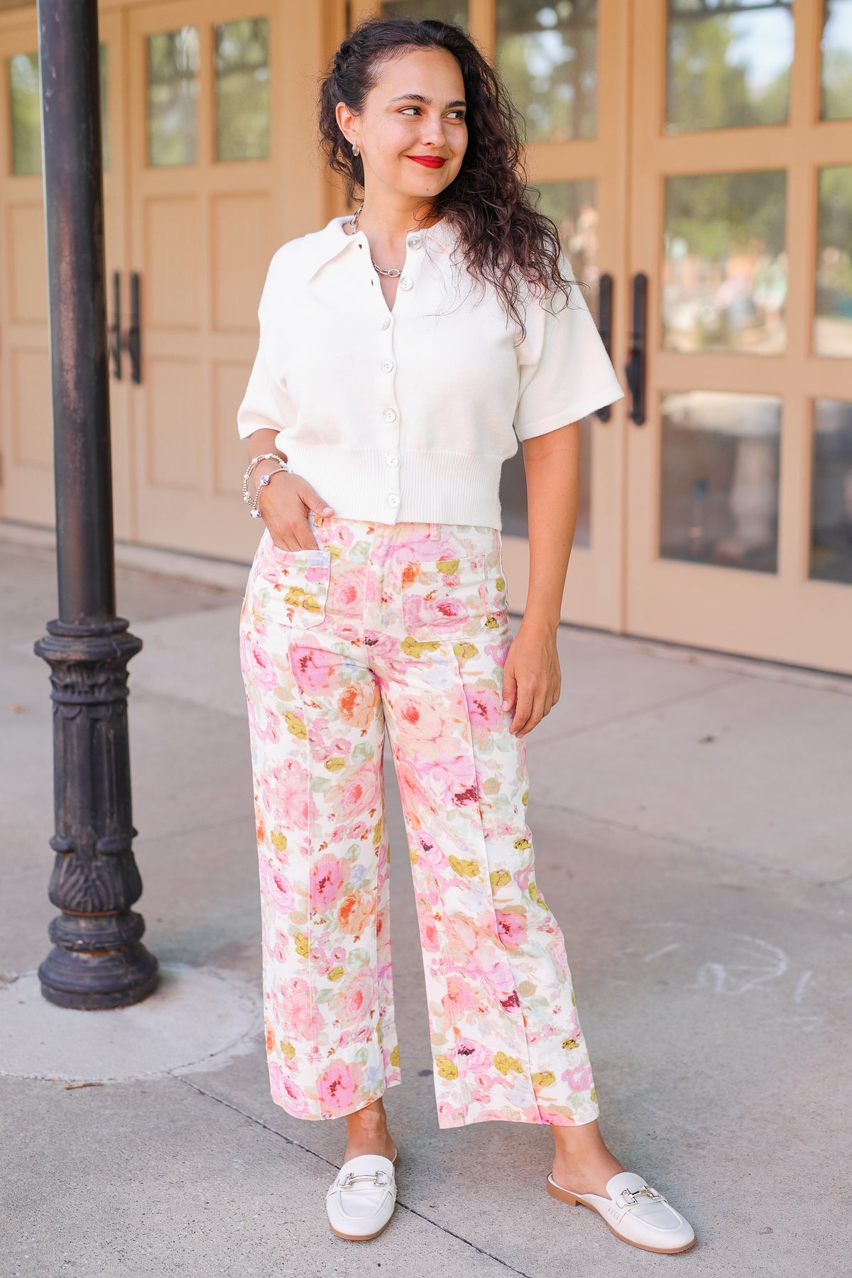 Brunette model standing outside wearing a off white cardigan and floral pattern denim pants.