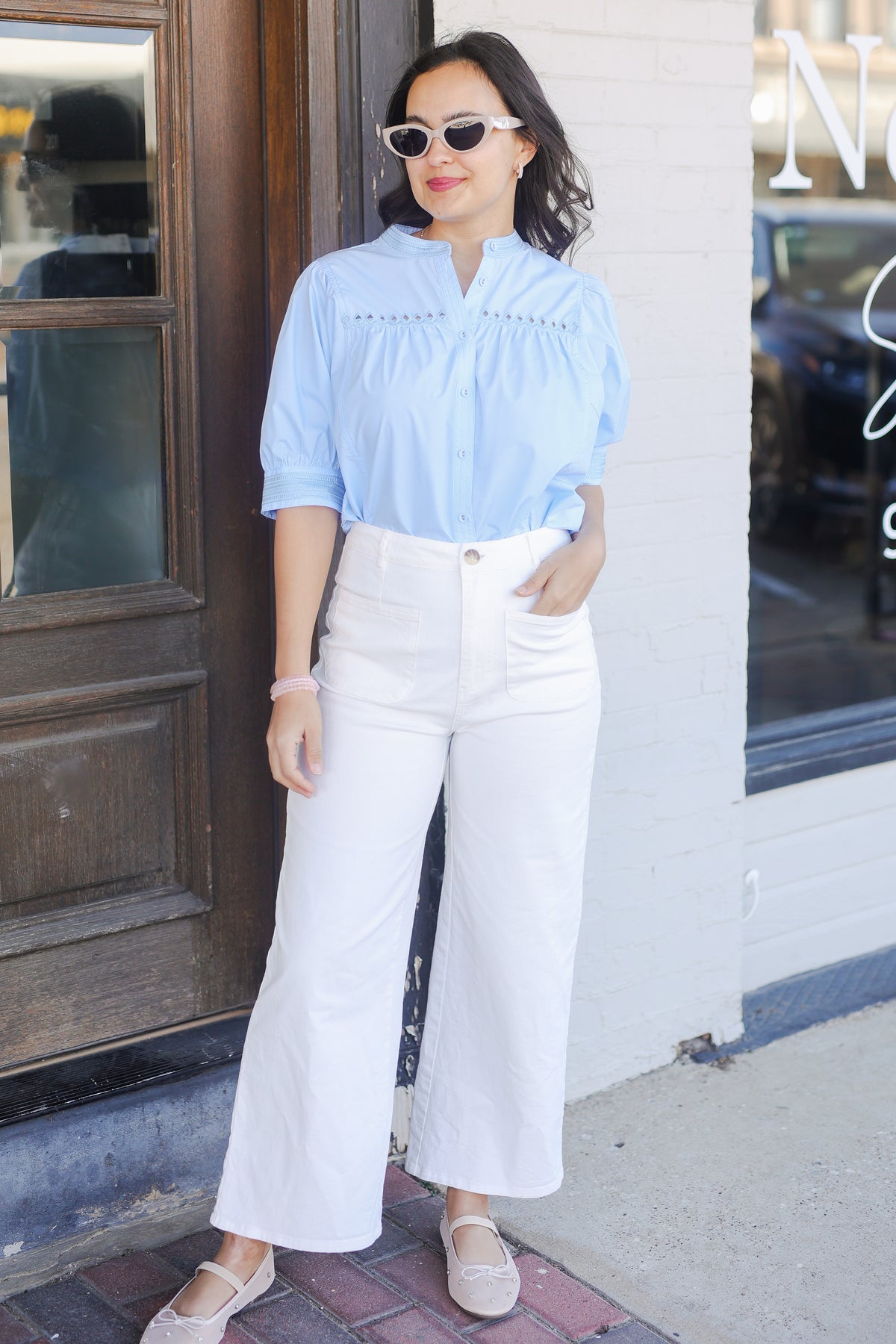 Woman wearing a light blue blouse and white pants outside with sunglasses.