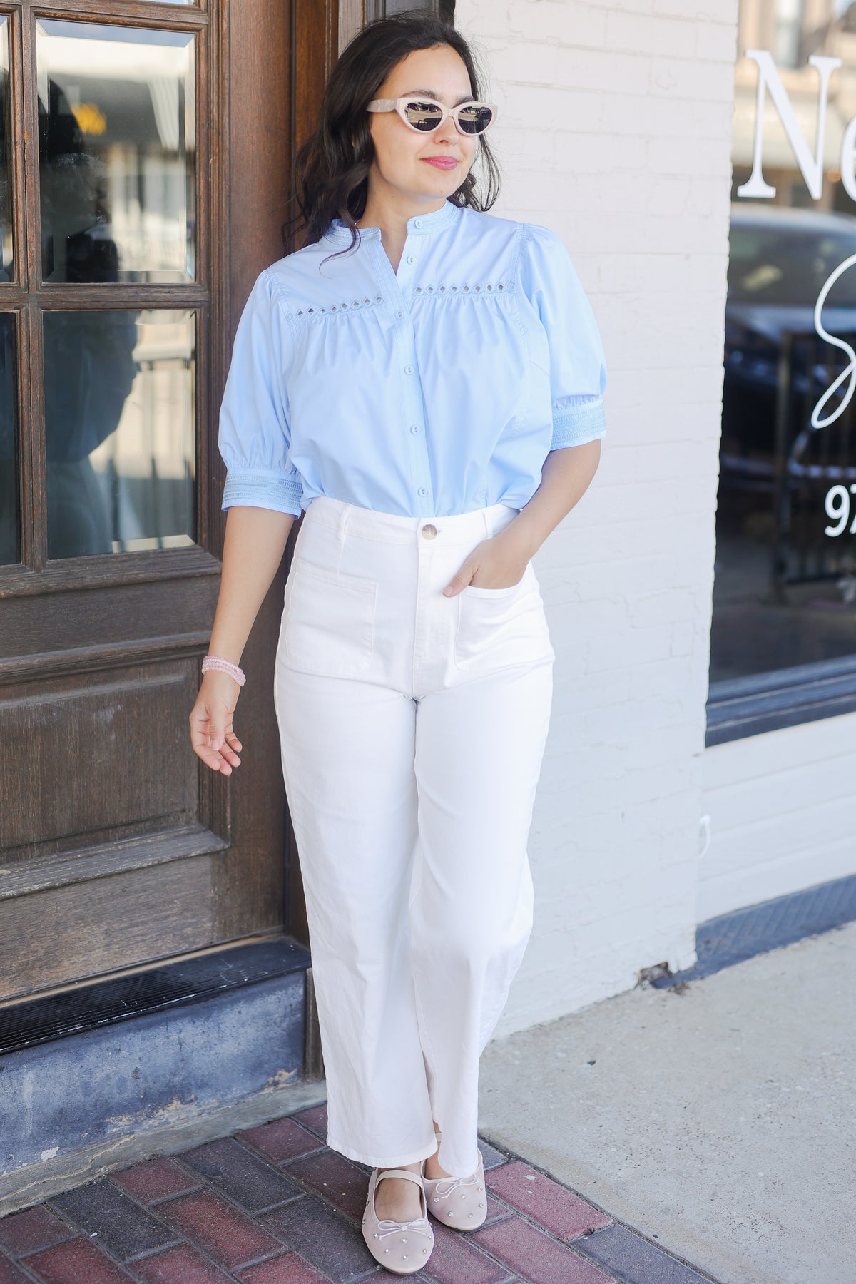 Woman wearing a light blue blouse and white pants outside with sunglasses.