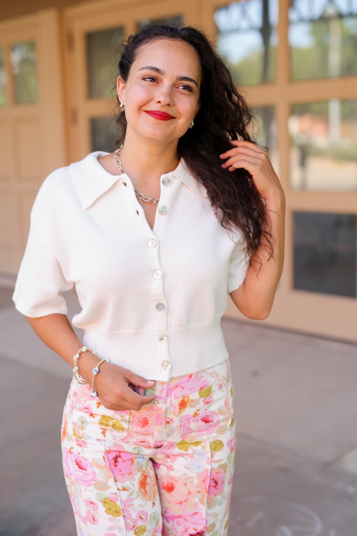 Brunette model standing outside wearing a off white cardigan and floral pattern denim pants.