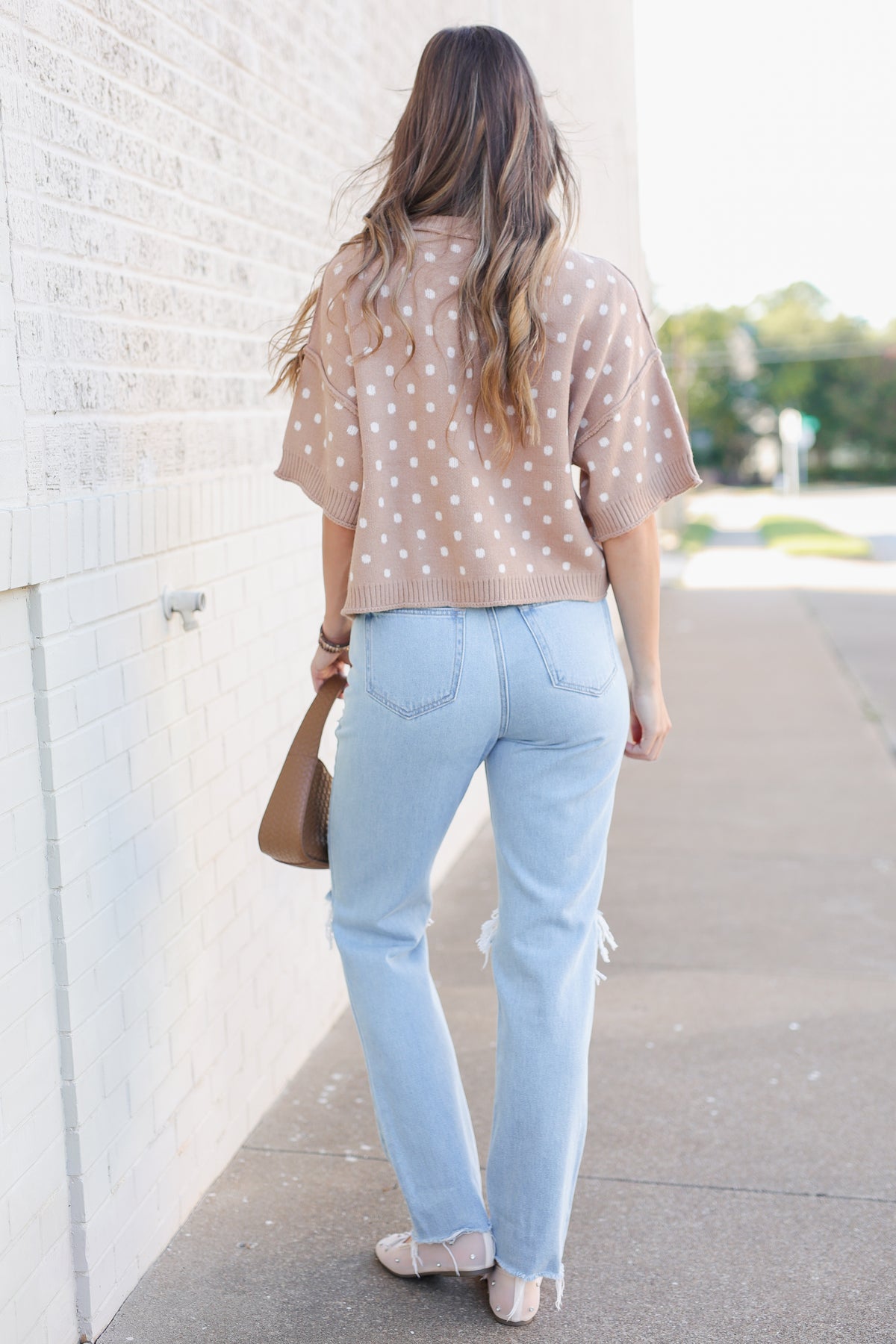 Blonde model standing outside near a brick wall wearing a camel colored sweater top with off white dots, blue jeans and flats.