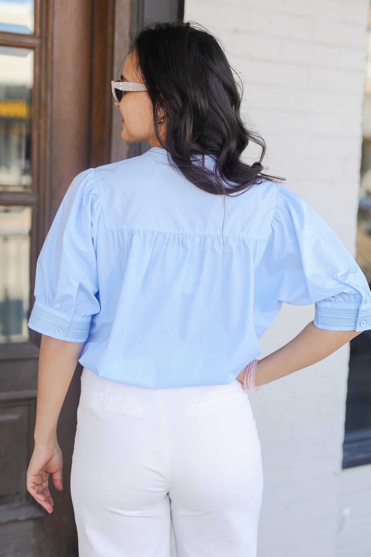 Woman wearing a light blue blouse and white pants outside with sunglasses.