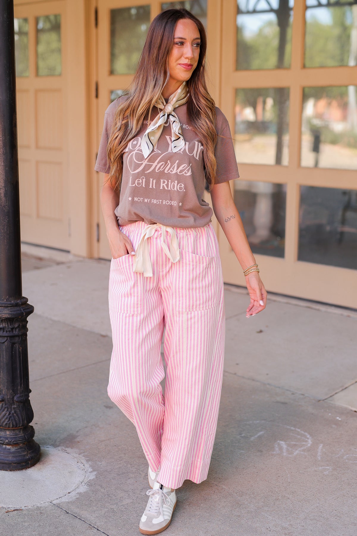 A woman stands on a sidewalk wearing a brown graphic T-shirt with white script text, pink and white vertically striped wide-leg pants, and white sneakers. The background shows beige building doors and windows.
