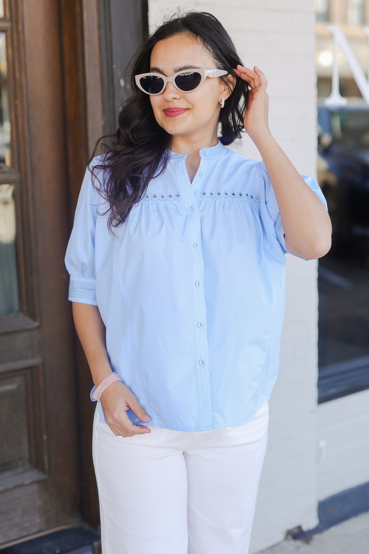 Woman wearing a light blue blouse and white pants outside with sunglasses.