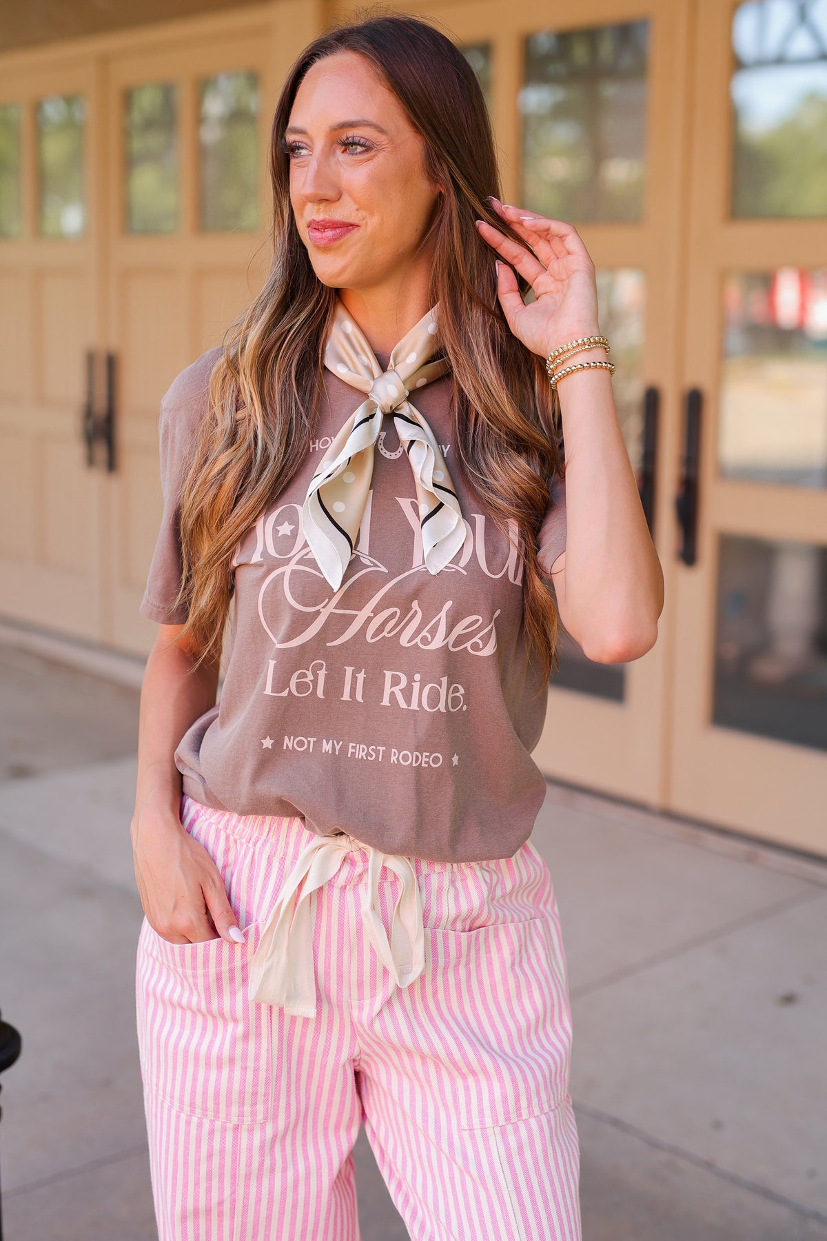 A woman wearing a brown graphic t-shirt with 'Horses Let It Ride' text, pink and white striped pants, and a cream neck scarf, standing outdoors in front of a beige building.