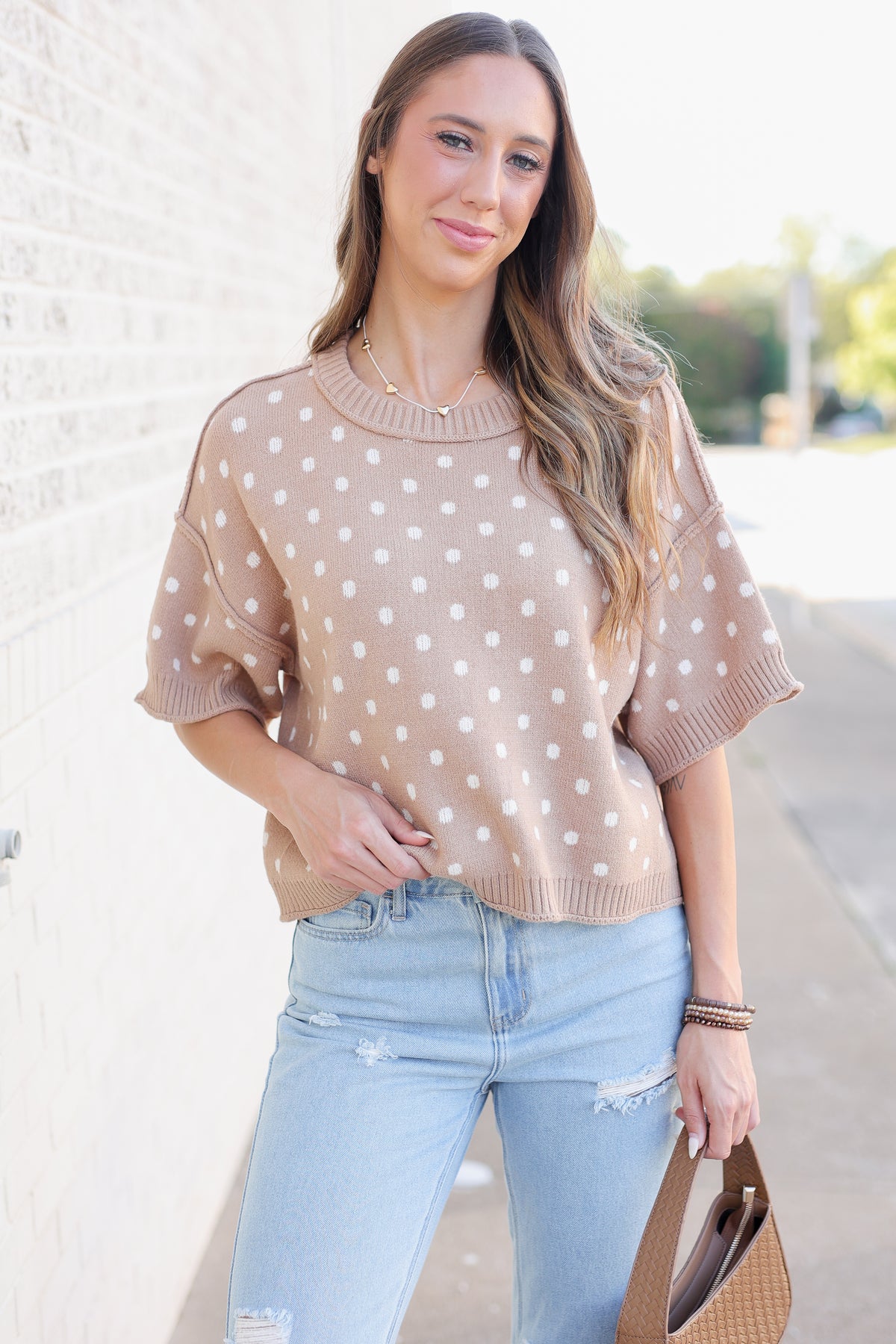 Blonde model standing outside near a brick wall wearing a camel colored sweater top with off white dots, blue jeans and flats.