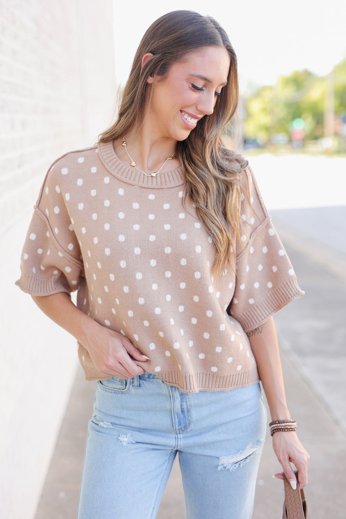 Blonde model standing outside near a brick wall wearing a camel colored sweater top with off white dots, blue jeans and flats.