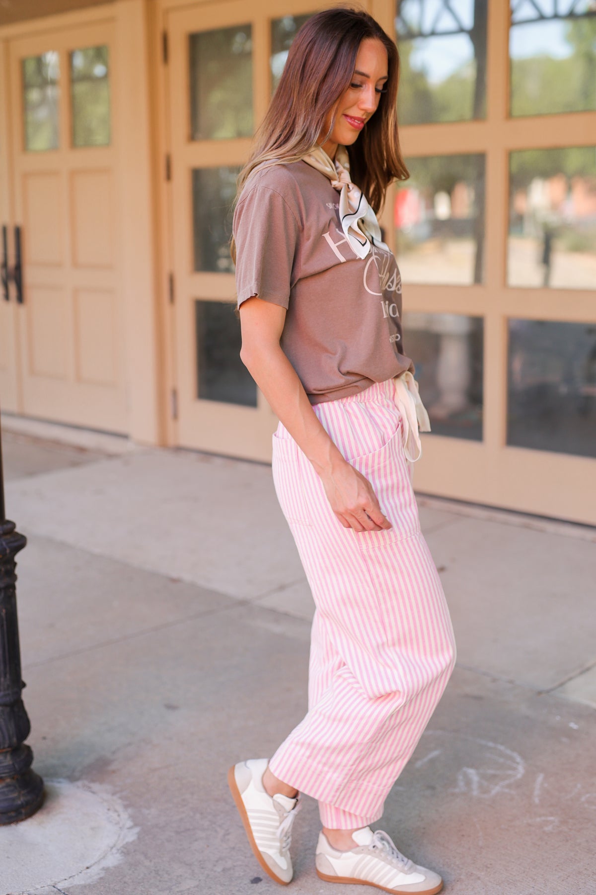 A woman wearing a brown graphic t-shirt, pink and white striped wide-leg pants, and white sneakers, standing on a sidewalk in front of a building with tan doors.