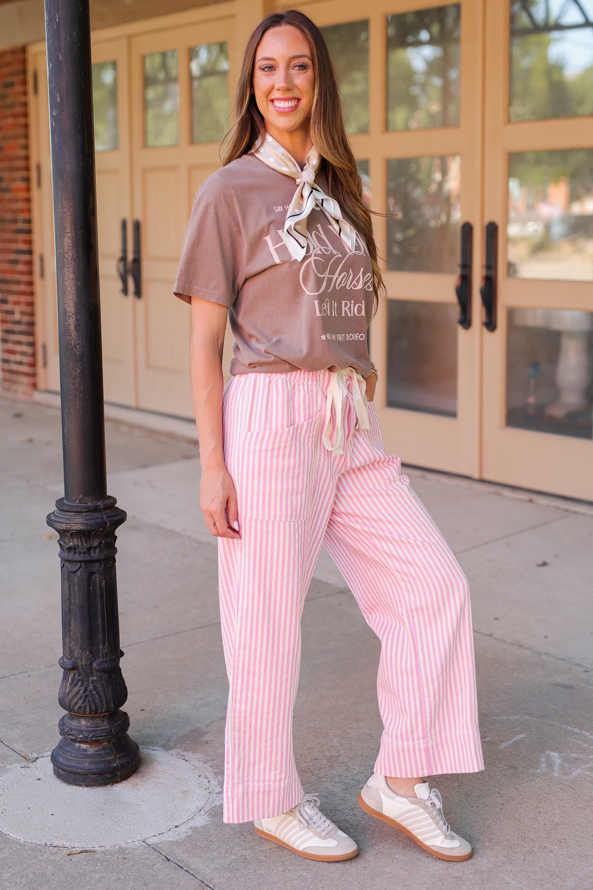 A woman stands outdoors in front of a building with glass doors, wearing a brown graphic T-shirt, pink and white vertically striped wide-leg pants, a cream neck scarf, and white sneakers.