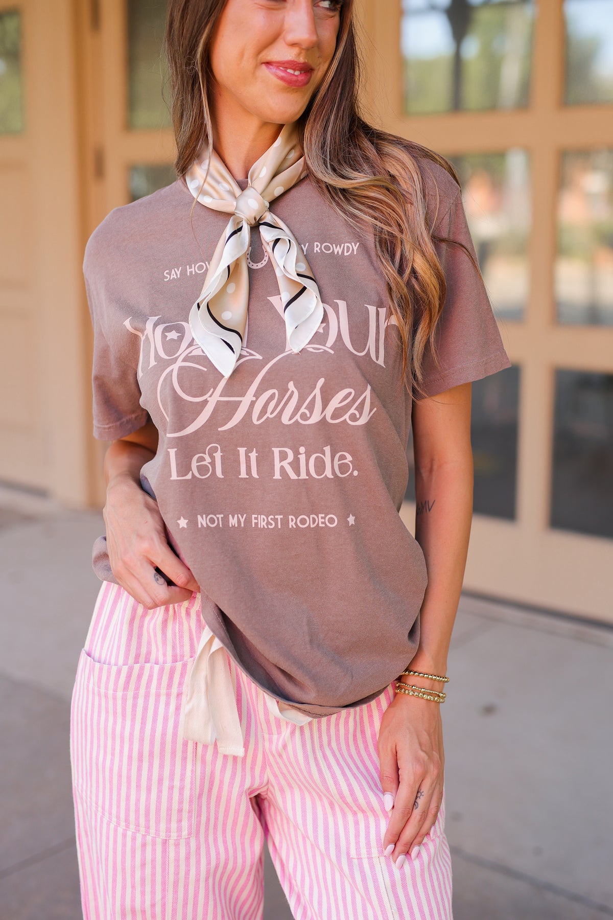 A woman wearing a brown graphic t-shirt with white text that reads 'Hold your Horses Let It Ride. NOT MY FIRST ROOSTER.