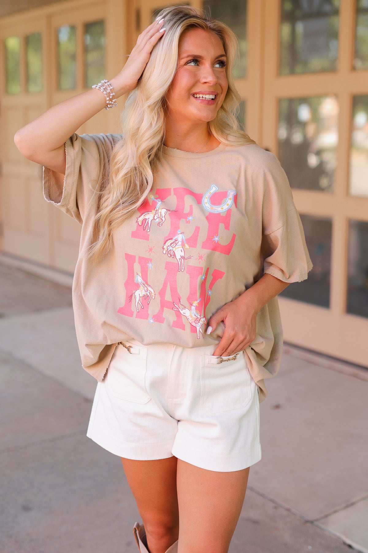 A woman wearing a beige graphic t-shirt with pink distressed lettering that reads 'SEE HAY' and white shorts, standing outdoors in front of a building with tan garage-style doors.