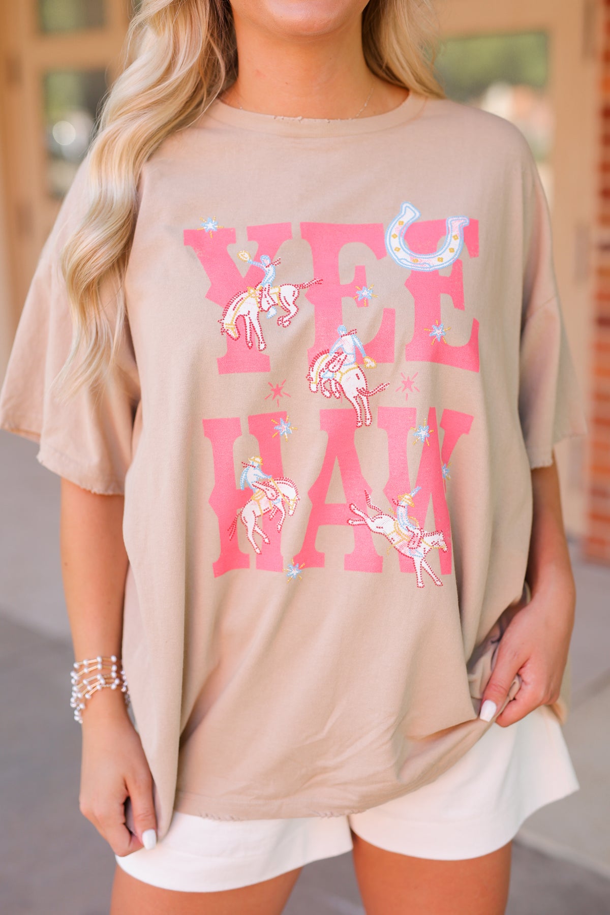 A woman wearing an oversized beige graphic T-shirt with pink 'YEE HAW' lettering and carousel horse illustrations, paired with white shorts.