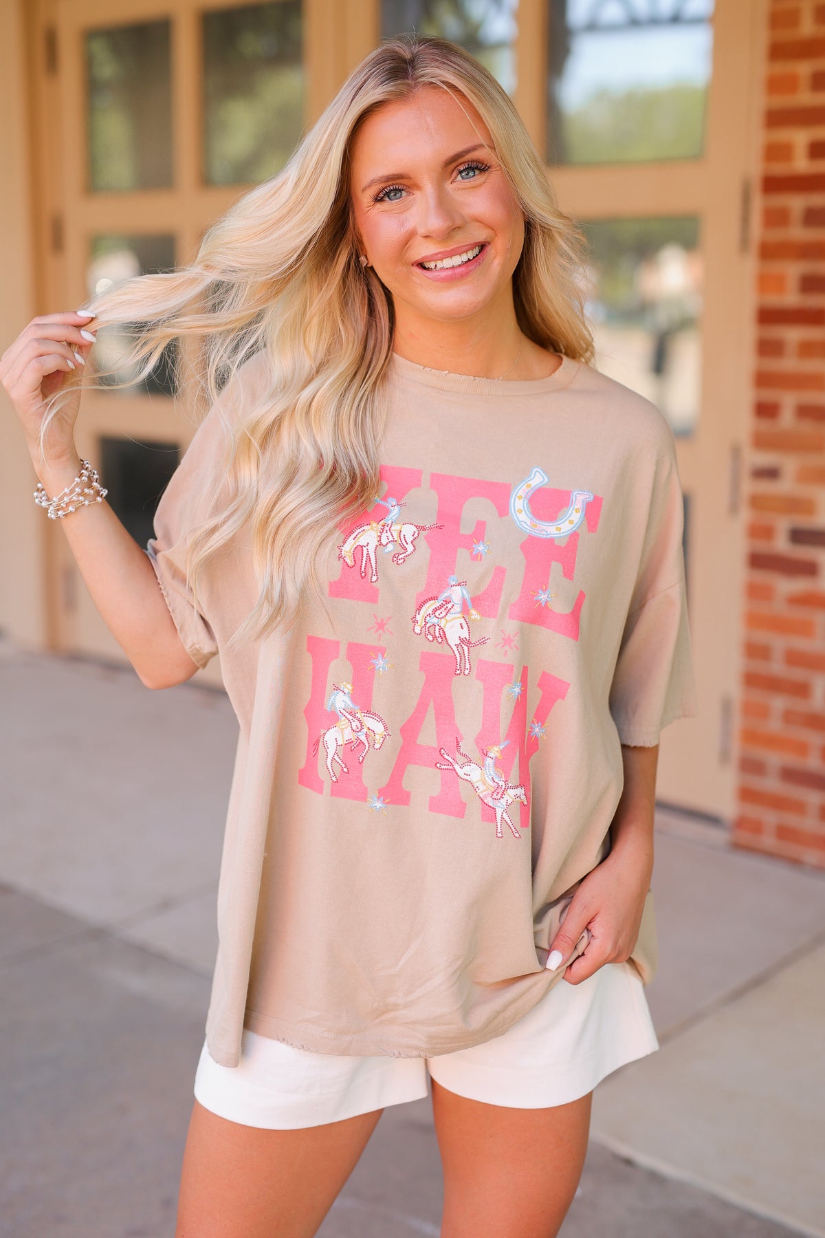 A woman wearing a beige oversized graphic t-shirt with pink and white text reading 'yee haw' and white shorts, standing outdoors in front of a building.