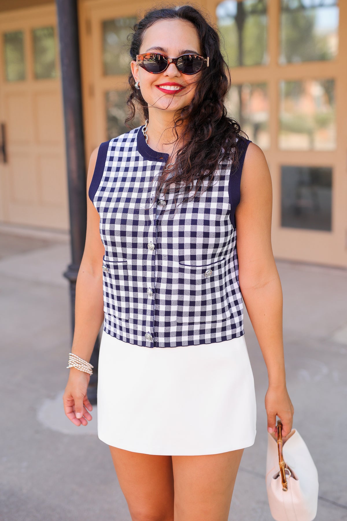 A woman wearing a sleeveless navy and white gingham button-front top with a white mini skirt, sunglasses, and holding a white hat, standing outdoors in front of a building with beige doors.
