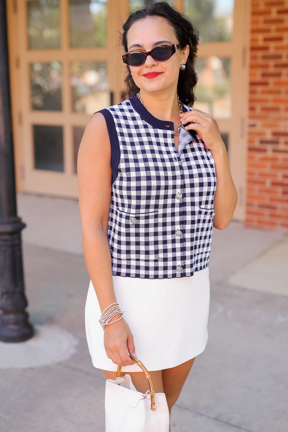 A woman wearing a navy and white gingham sleeveless vest with a white mini skirt, sunglasses, and a white handbag, standing outdoors in front of a building with brick and glass doors.