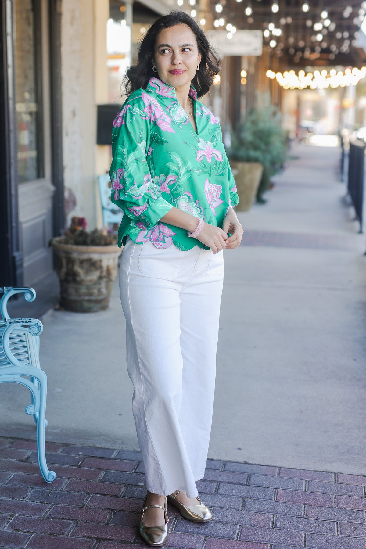 Woman wearing a green floral blouse and white pants standing on a sidewalk.
