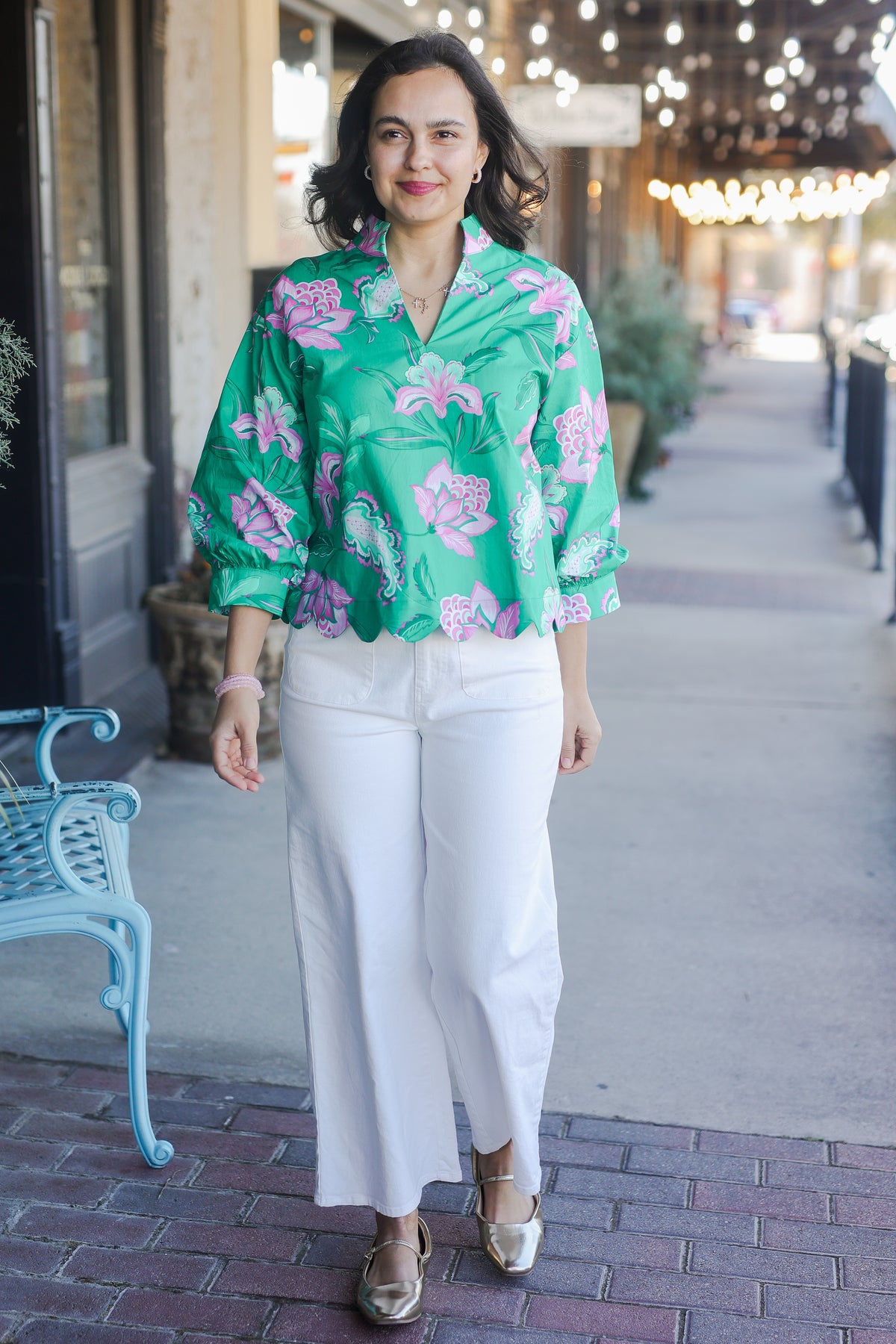 Woman wearing a green floral blouse and white pants on a sidewalk.