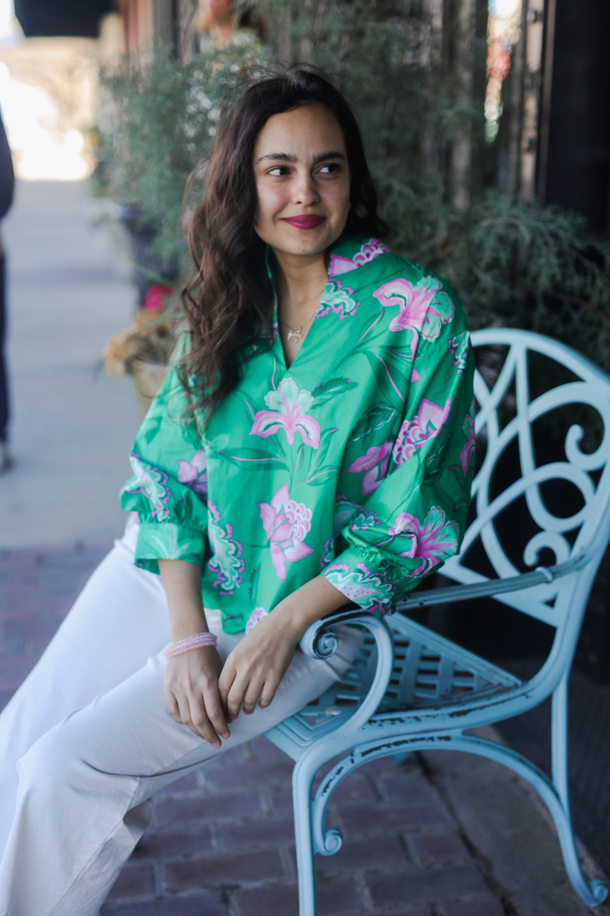 Woman in a green floral blouse sitting on a white chair outdoors.