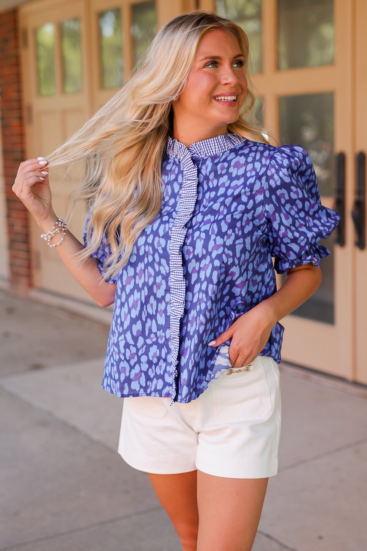 A woman wearing a blue and purple patterned puff-sleeve blouse with a striped collar and white shorts, standing outdoors in front of a building with tan doors.