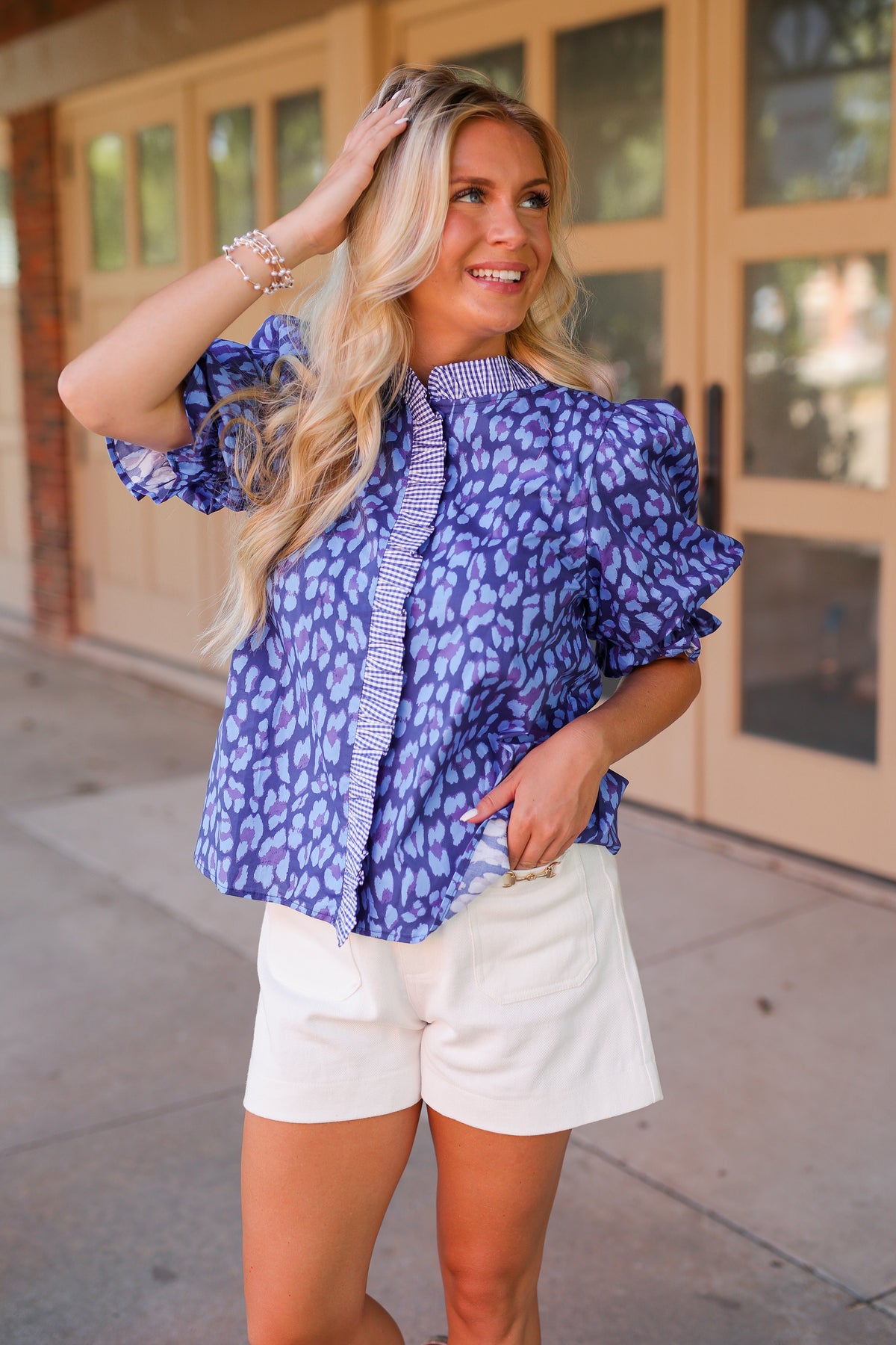 A woman posing outdoors in front of a building, wearing a blue and purple patterned puff-sleeve blouse with a striped tie-neck detail and white shorts.