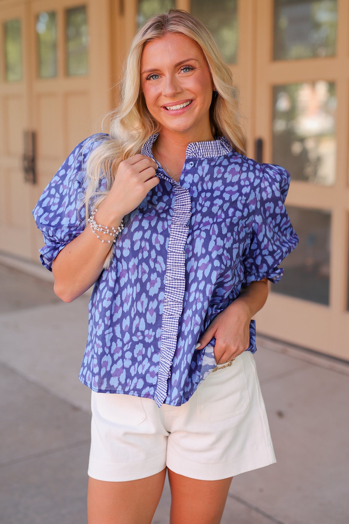 A woman modeling a blue and purple leopard-print blouse with puff sleeves and a striped placket, paired with white shorts, standing outdoors in front of a beige building.