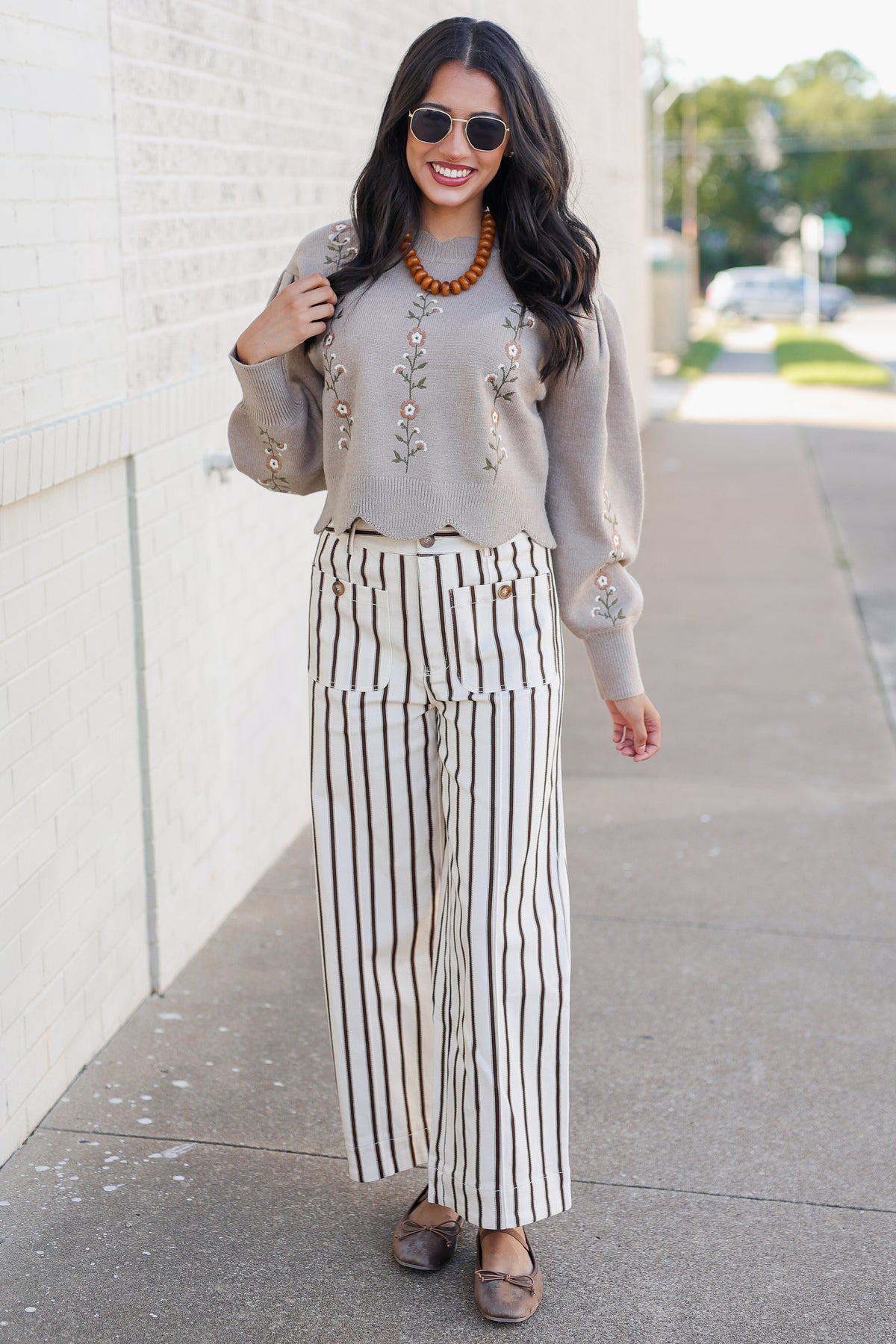 A woman wearing a beige knit sweater with floral embroidery, cream and black striped wide-leg pants, and brown flat shoes, standing on a sidewalk next to a white brick wall.