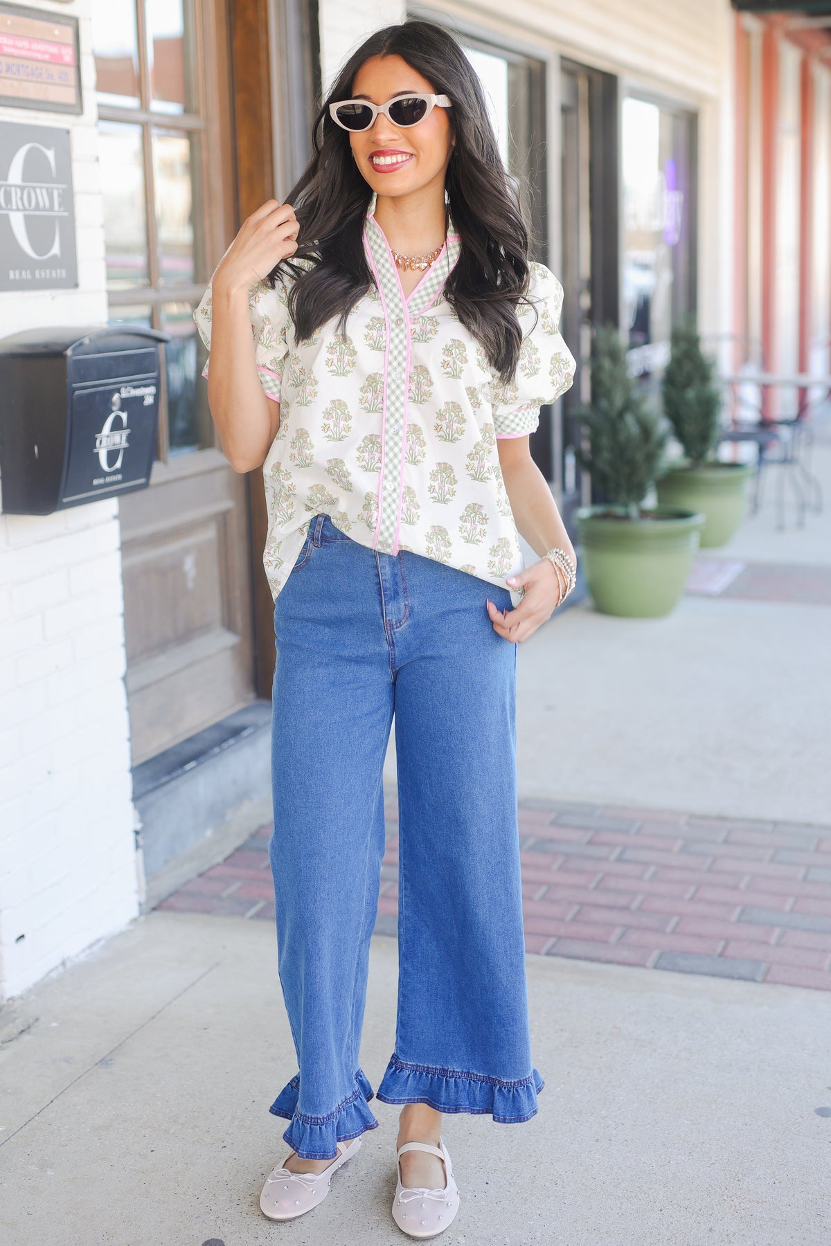 Woman wearing a floral blouse and blue jeans standing on a sidewalk.