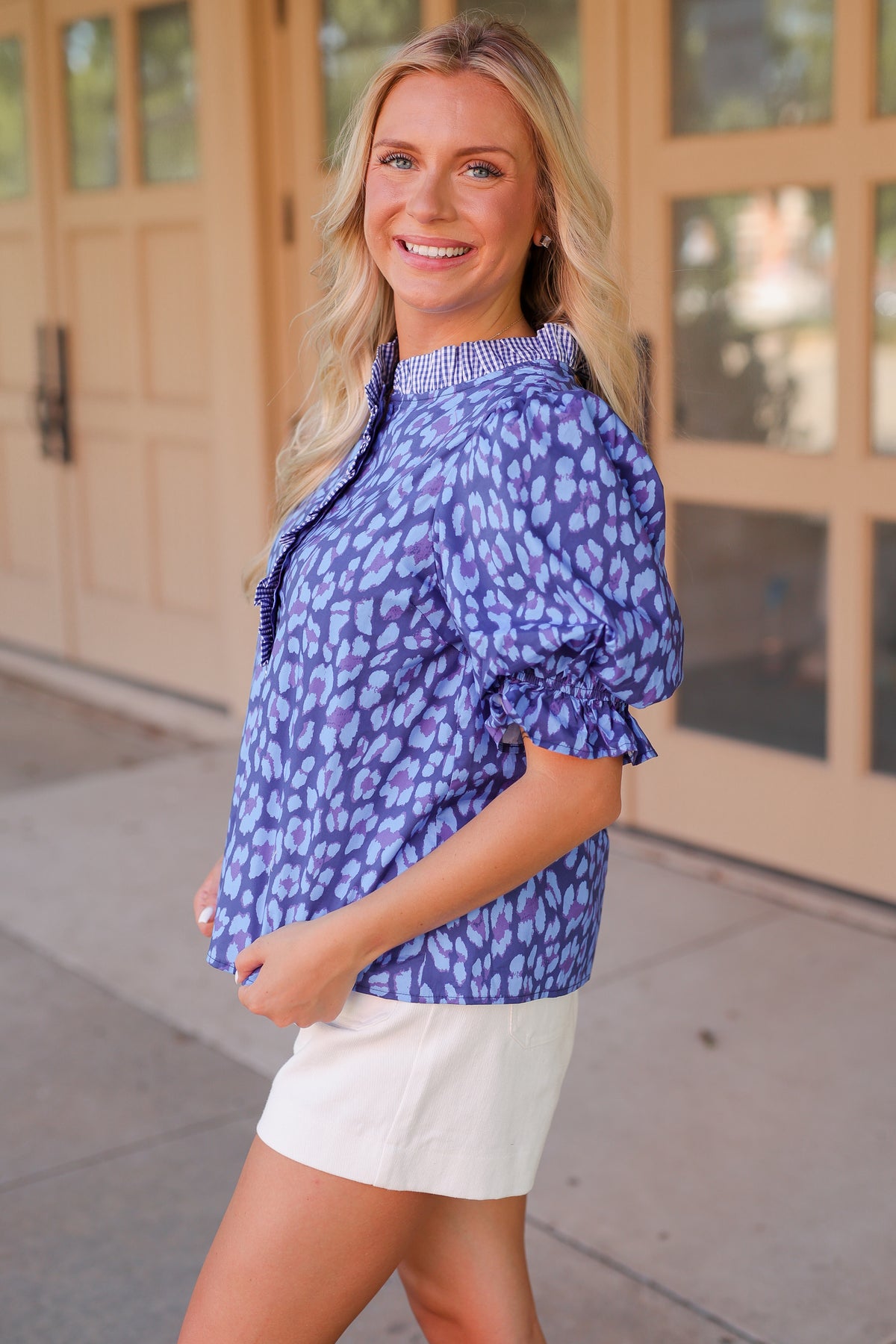 A woman wearing a blue and purple leopard-print blouse with puff sleeves and white shorts, standing outdoors in front of a beige building.