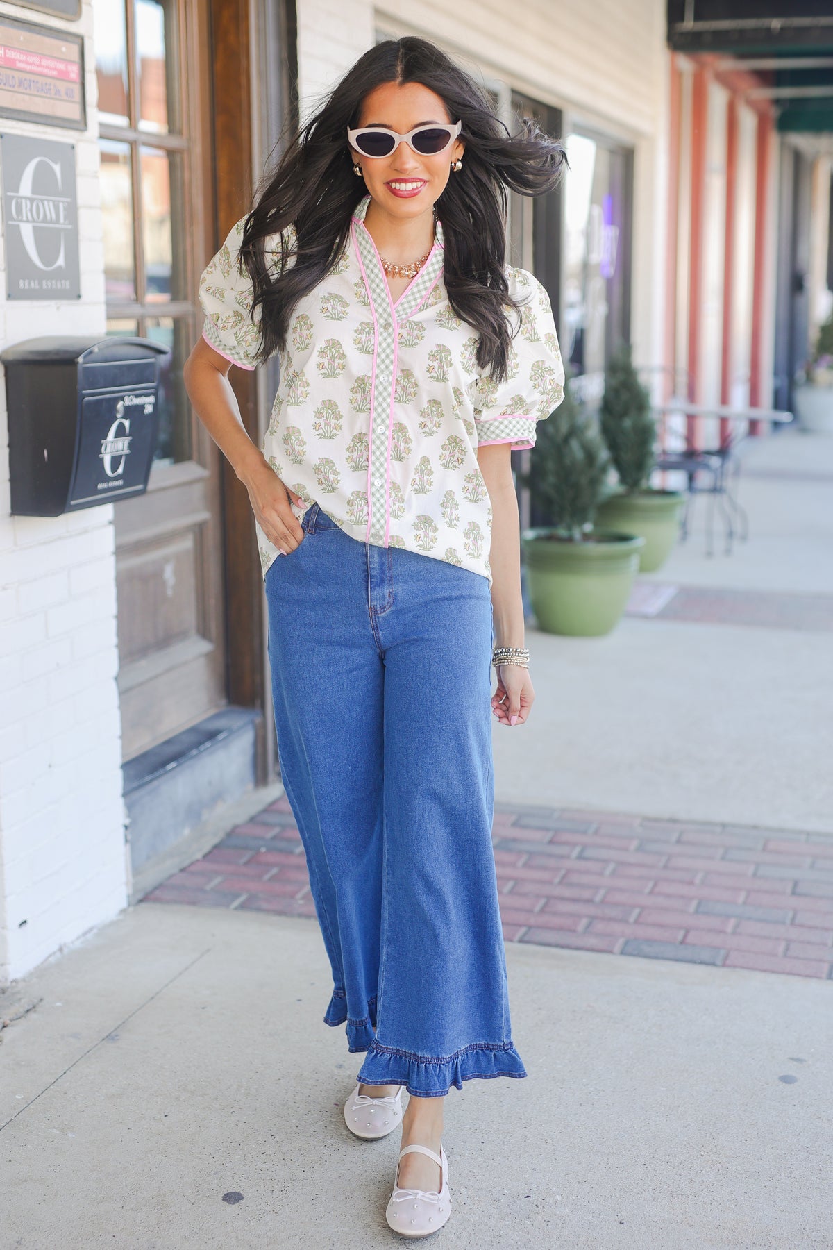 Woman wearing a floral blouse and blue jeans standing on a sidewalk.