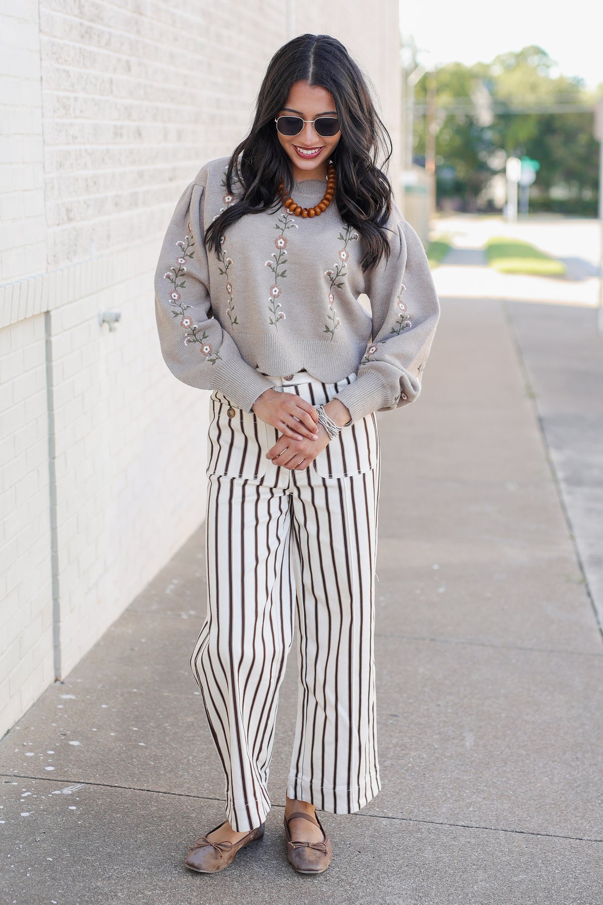 A woman stands on a sidewalk wearing a light beige crewneck sweater with floral embroidery, paired with black and white vertically striped wide-leg pants and brown flat shoes.