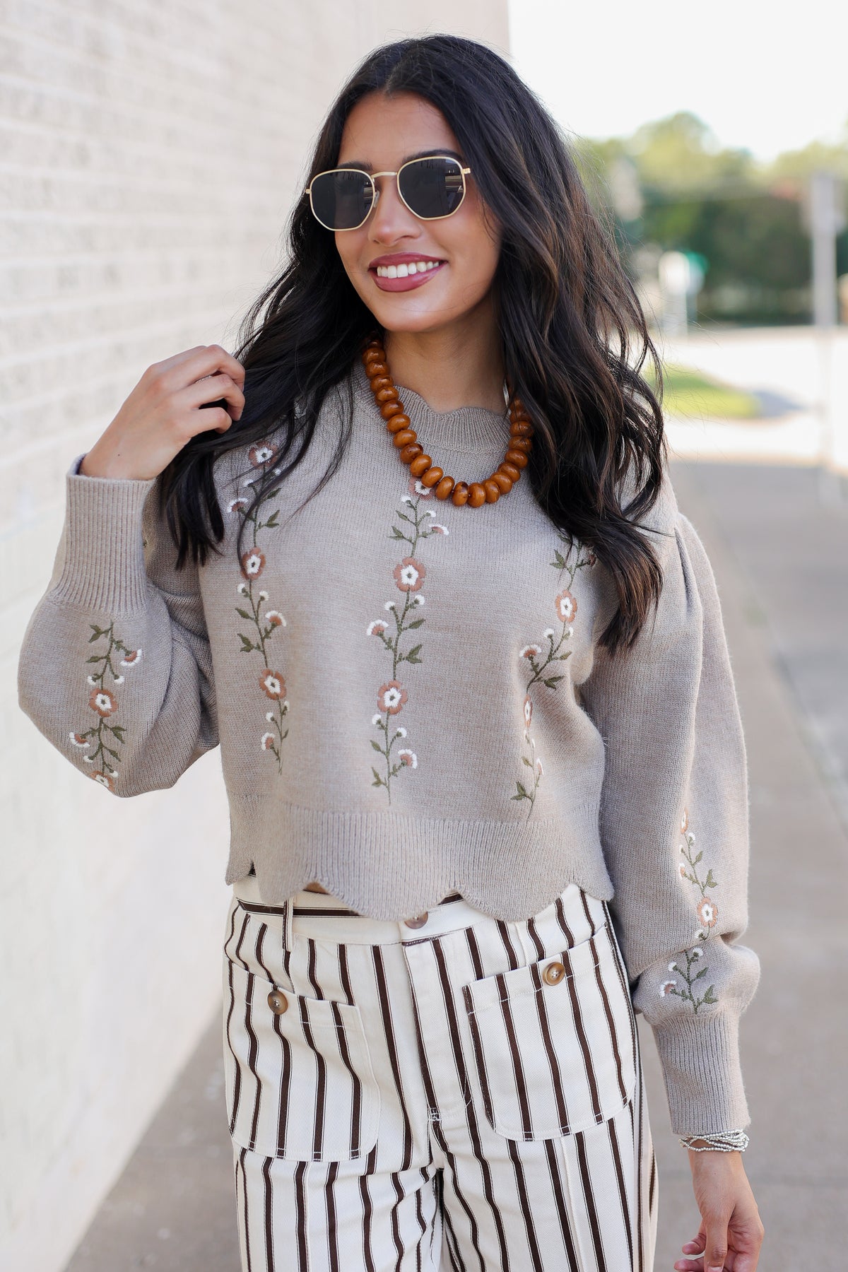 A woman wearing a light beige knit sweater with floral embroidery, paired with black and white vertically striped pants, a brown beaded necklace, and sunglasses, standing outdoors against a light brick wall.