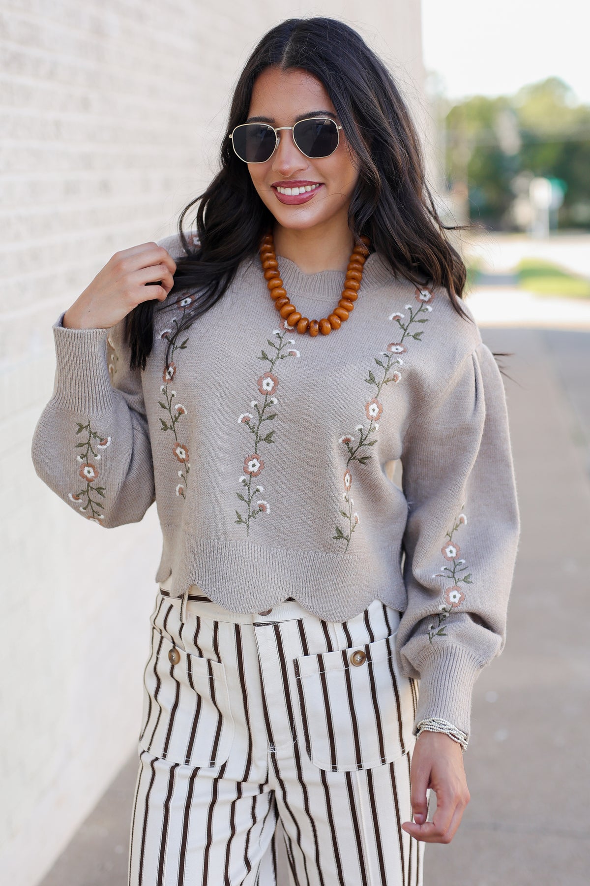 A woman wearing a light taupe knit sweater with floral embroidery, paired with black and white vertically striped pants, a wooden bead necklace, and sunglasses, standing outdoors against a light brick wall.