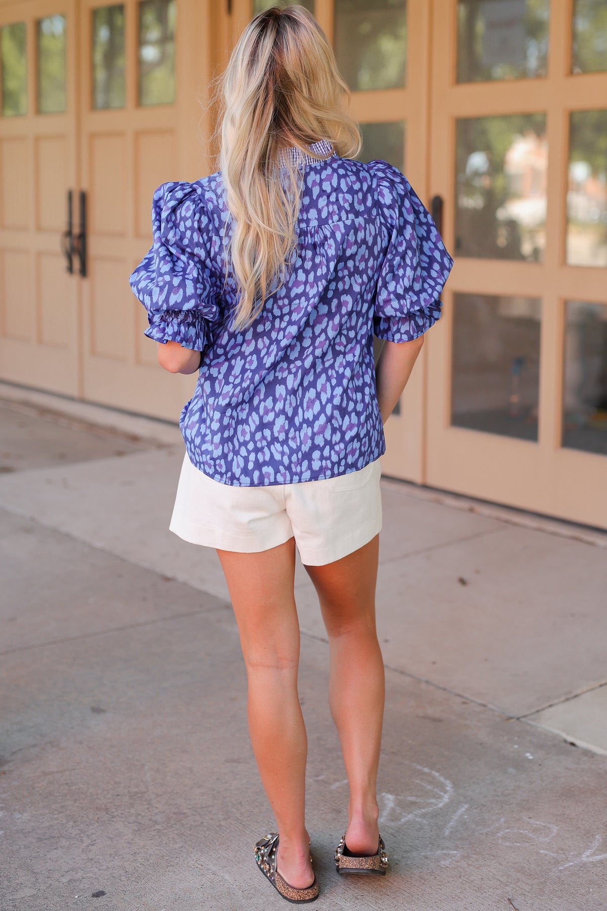 A woman viewed from behind wearing a blue and white patterned puff-sleeve blouse, cream shorts, and brown sandals, standing on a sidewalk in front of beige building doors.