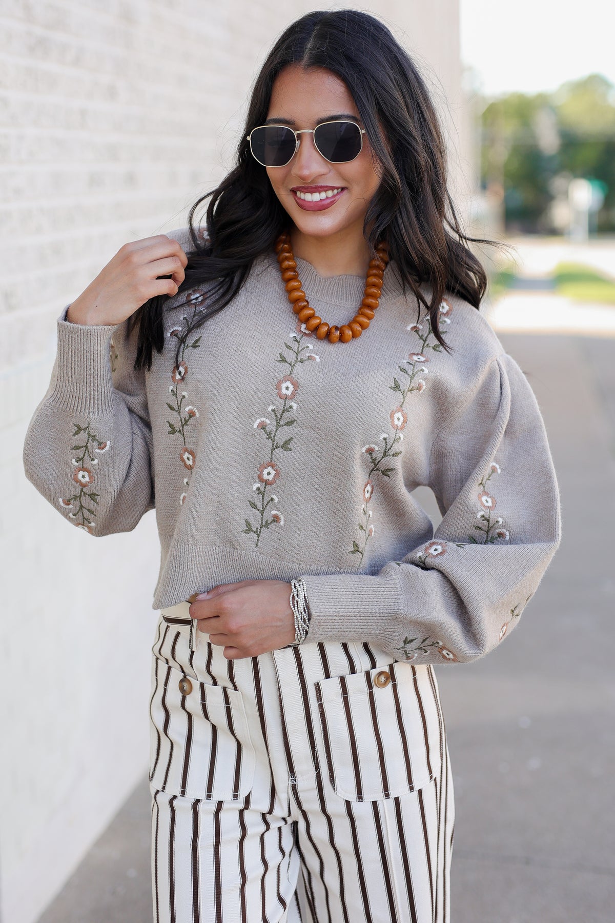 A woman wearing a light gray knit sweater with vertical floral embroidery, paired with black and white vertically striped pants, a wooden bead necklace, and sunglasses, standing outdoors against a light brick wall.