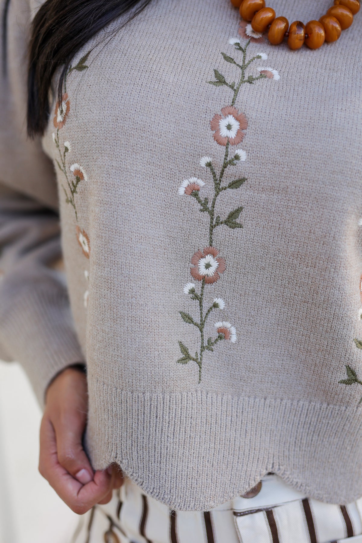 Close-up of a person wearing a beige knit sweater with vertical floral embroidery in pink, white, and green, paired with a chunky brown wooden bead necklace.