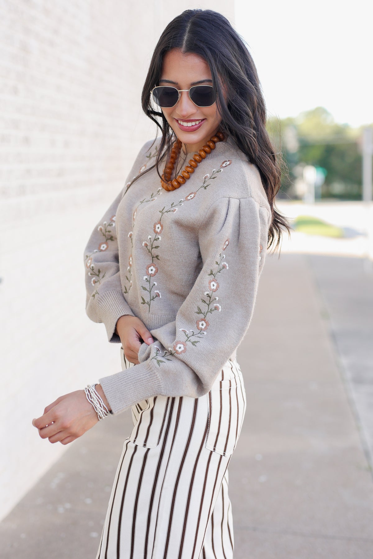 A woman wearing a light beige knit sweater with floral embroidery and puffed sleeves, paired with a cream and brown vertically striped skirt, sunglasses, and a wooden beaded necklace, standing outdoors on a sidewalk.