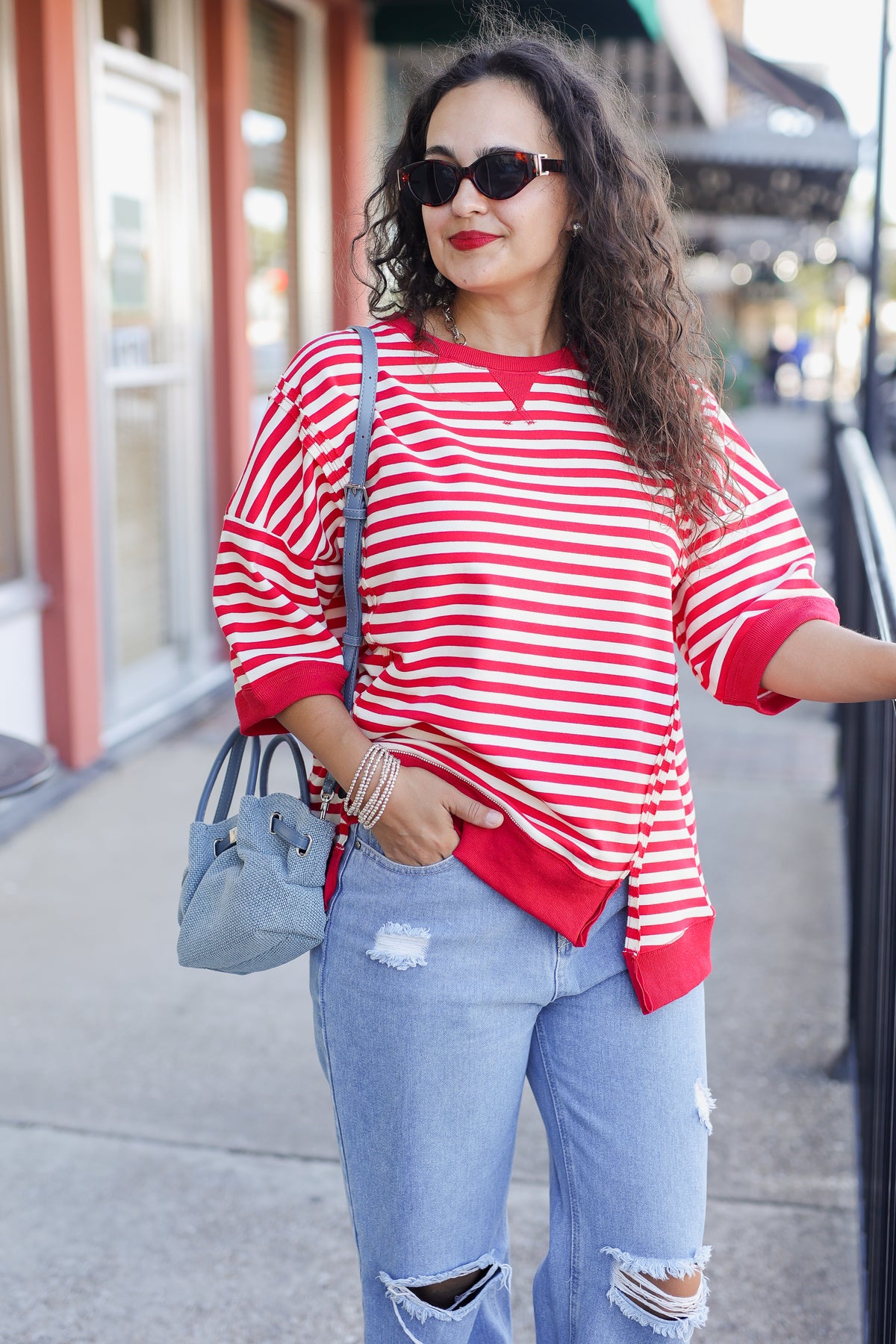 Woman wearing a red and white striped shirt and ripped jeans, standing on a sidewalk.