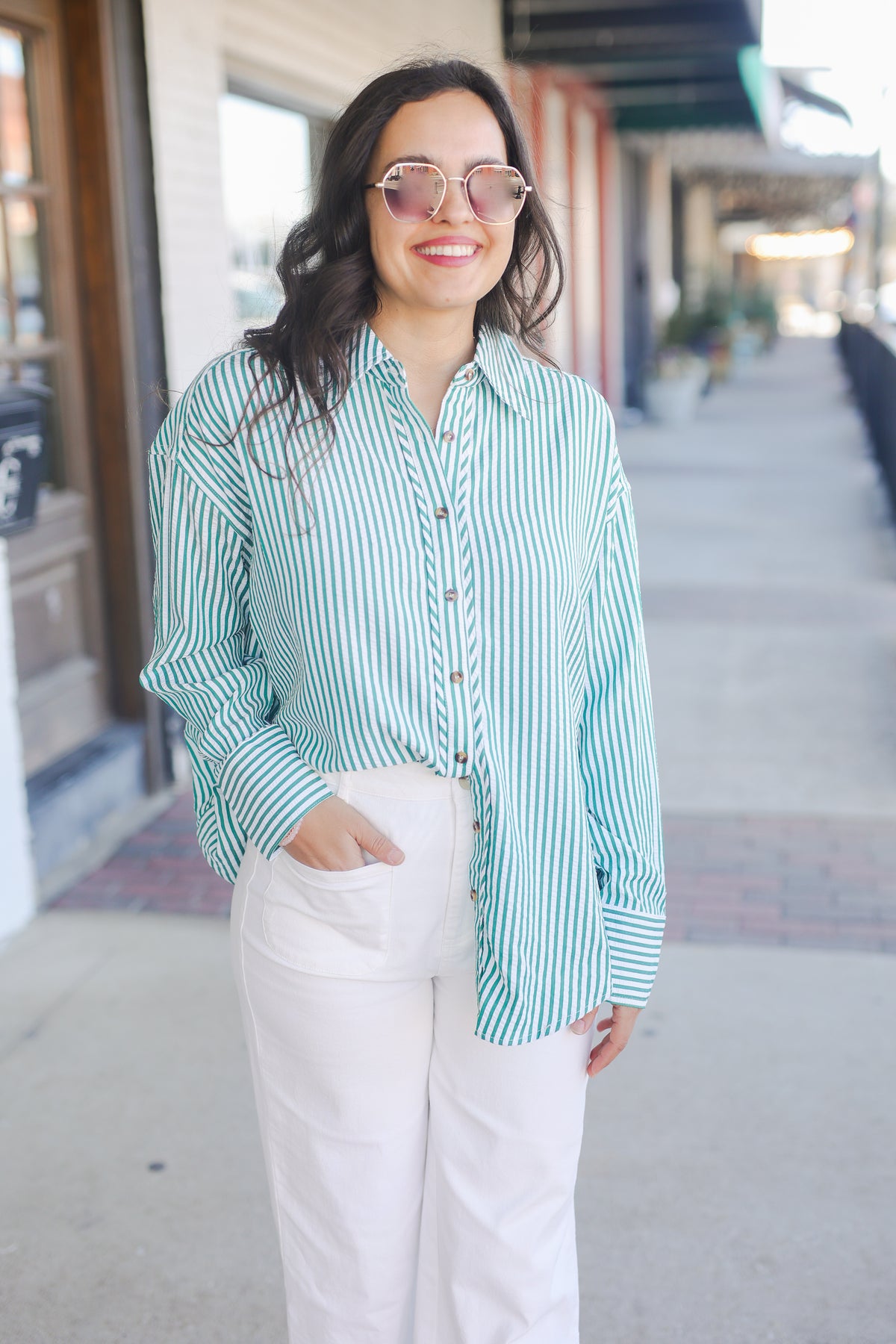 Woman wearing a green and white striped shirt and white pants standing on a sidewalk. The Carolina Coast Stripe Button Down - Green