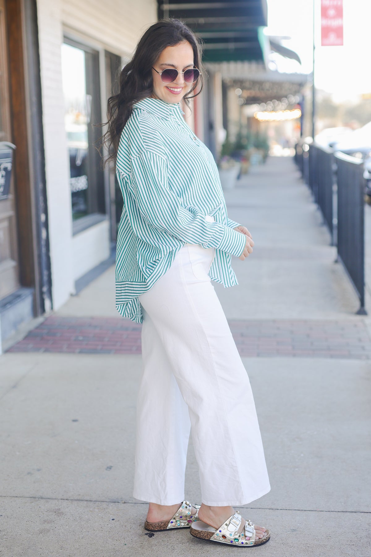 Woman wearing a green and white striped shirt and white pants on a sidewalk. The Carolina Coast Stripe Button Down - Green