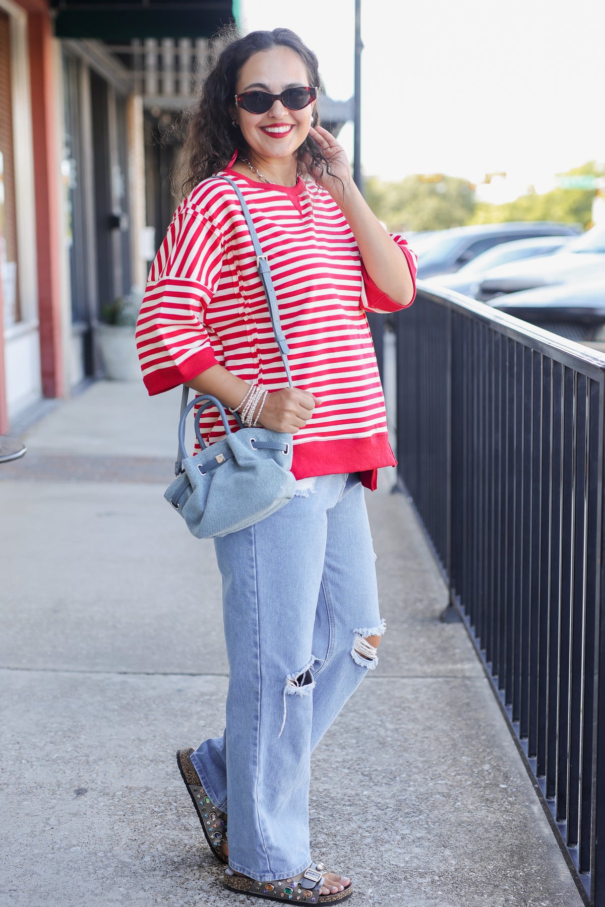 Woman wearing a red and white striped shirt and blue jeans, standing on a sidewalk.