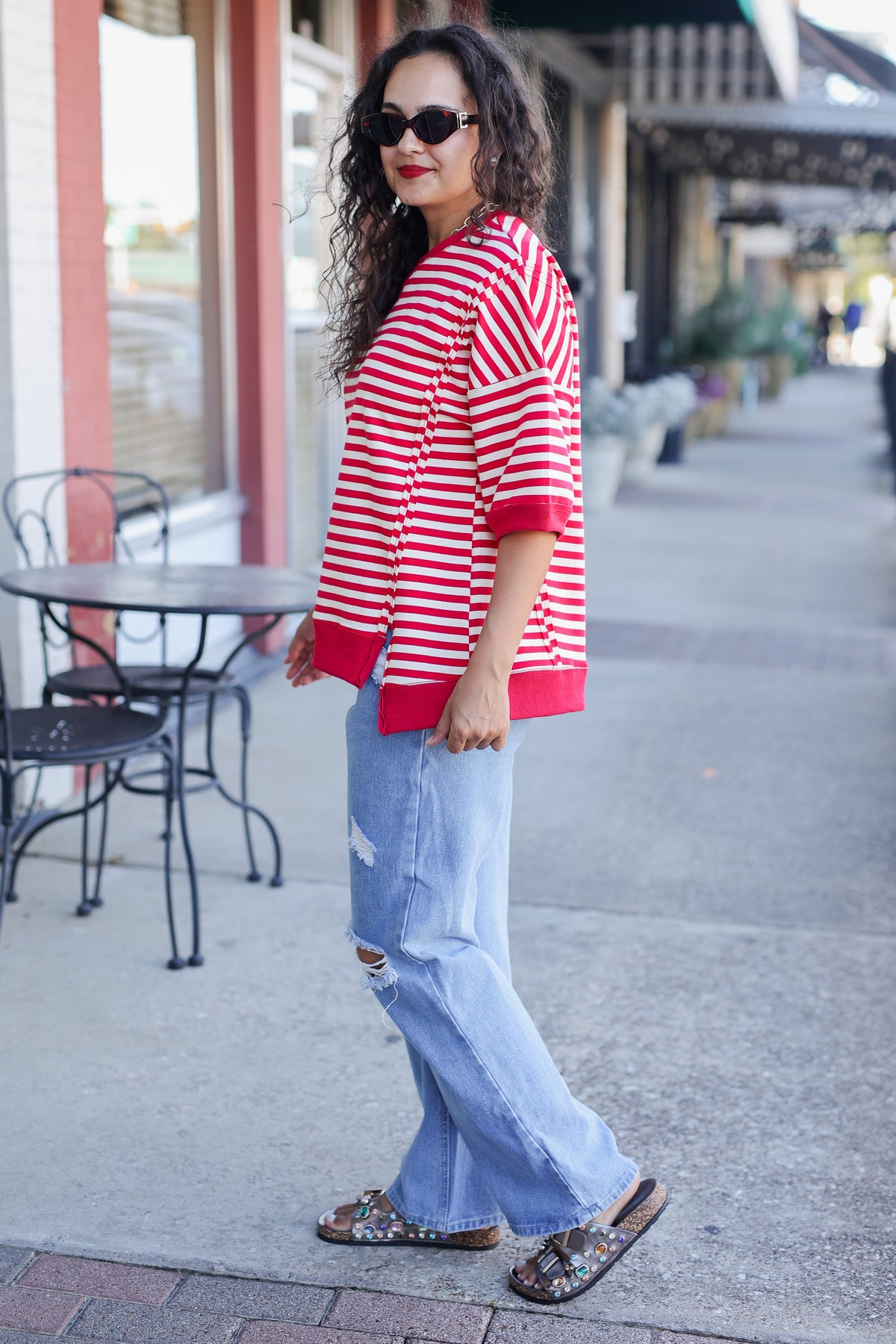Woman wearing a red and white striped shirt and light blue jeans standing on a sidewalk.