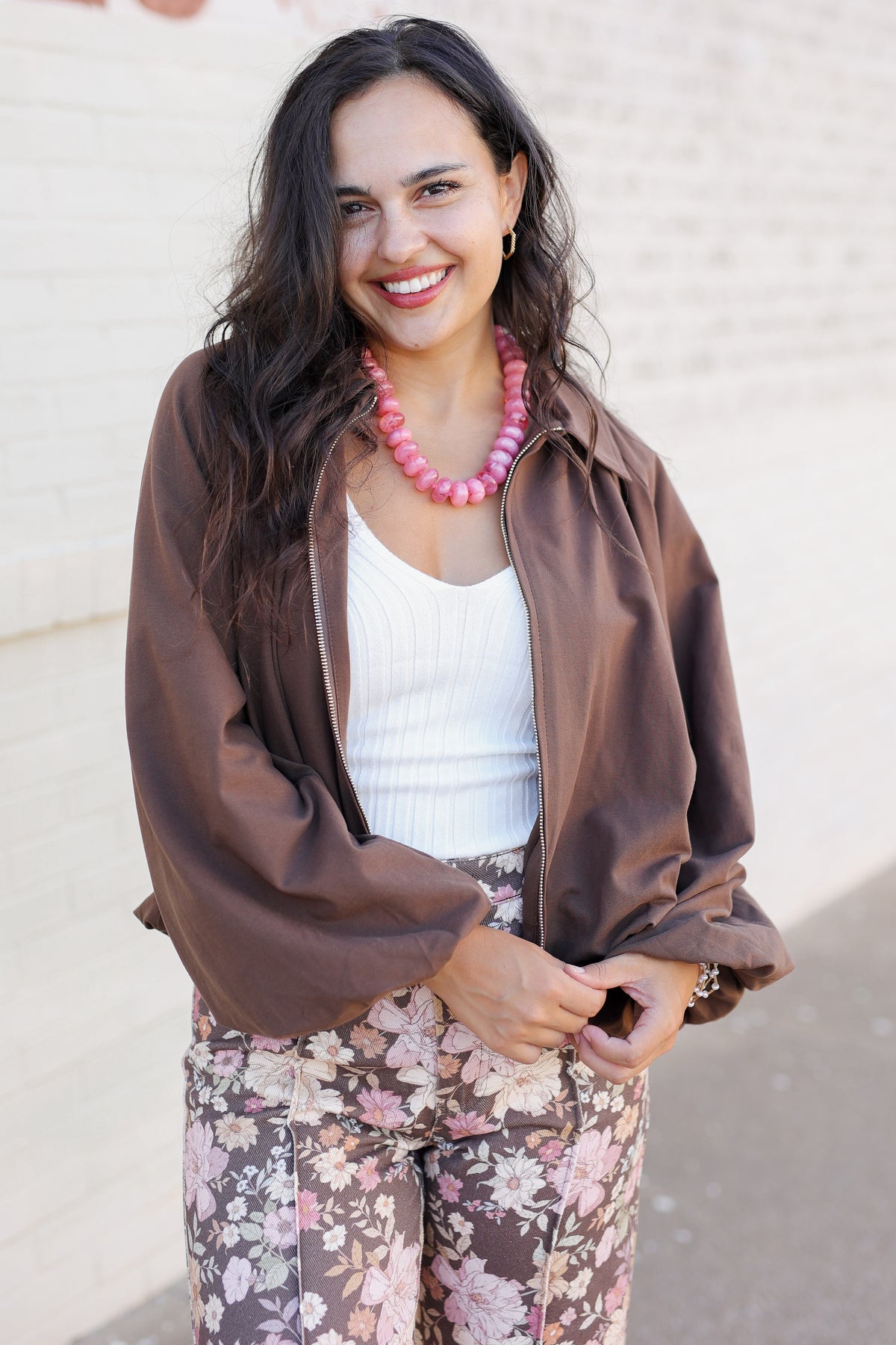A woman smiling and wearing a brown satin-like bomber jacket over a white ribbed tank top, paired with floral pants and a chunky pink beaded necklace, standing against a light brick wall.