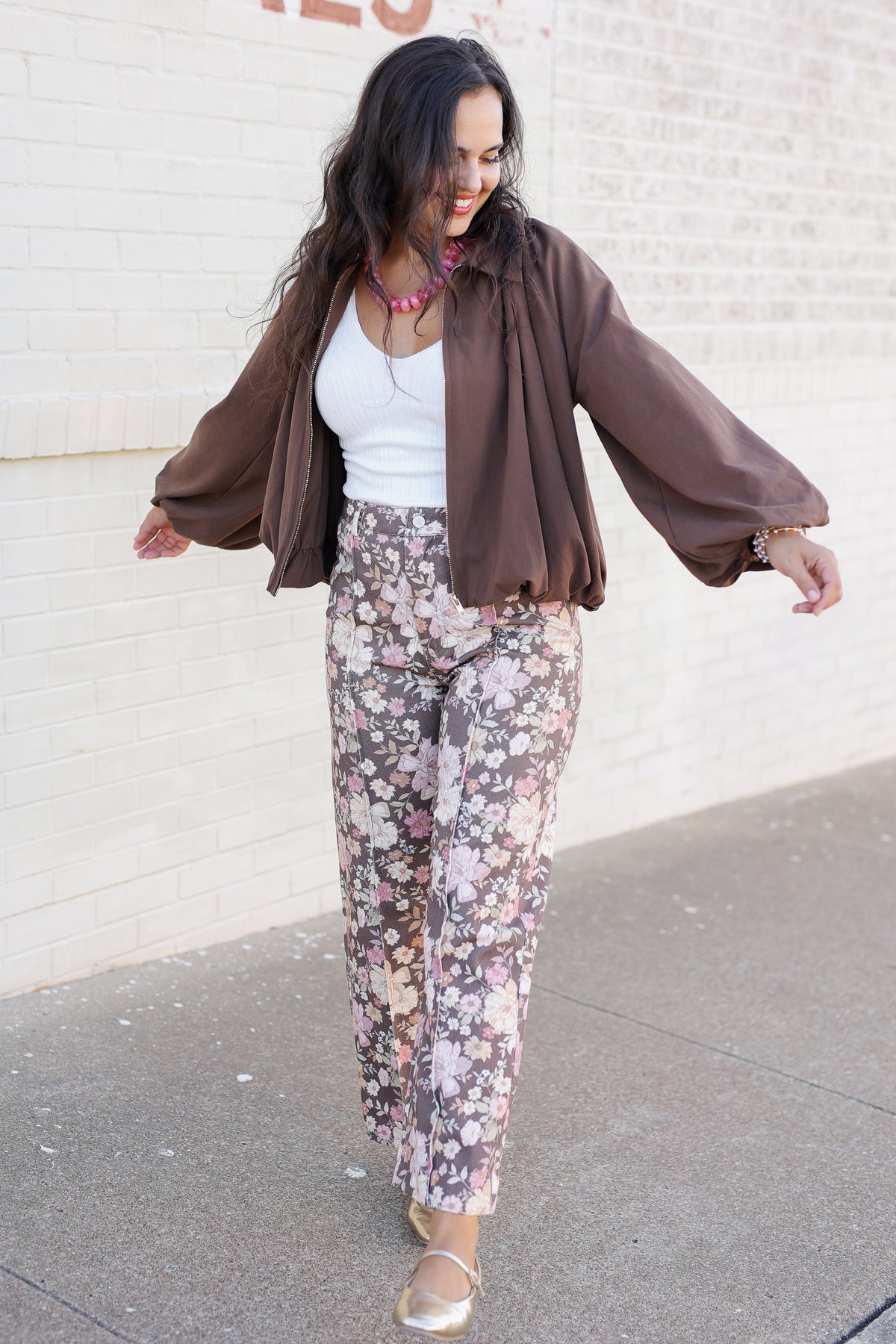 A woman modeling a brown sheer long-sleeve bomber jacket over a white tank top and floral print wide-leg pants, walking outdoors against a white brick wall.