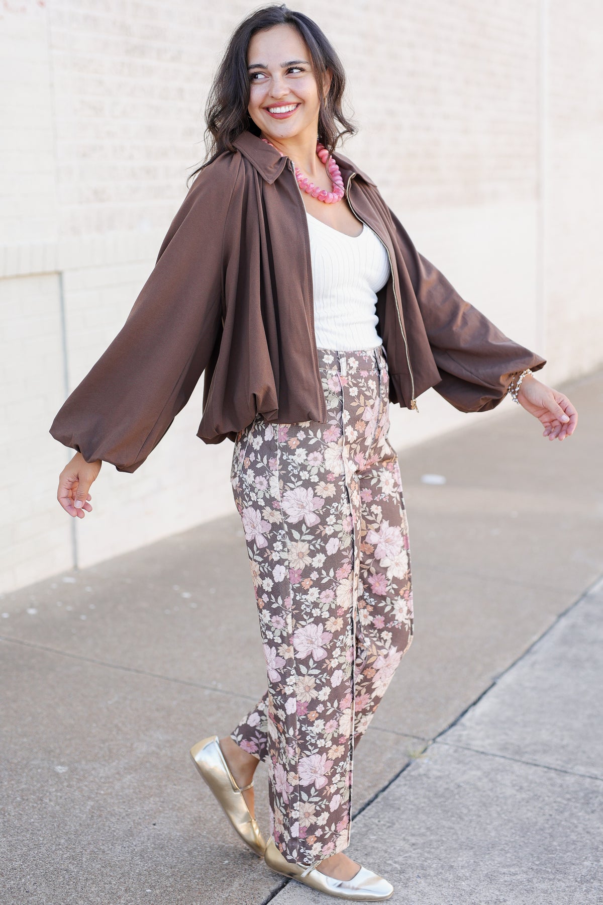A woman posing outdoors in a stylish outfit featuring a brown satin-like open-front jacket, a white top, and floral print pants in pink, beige, and brown tones, paired with metallic gold flats.