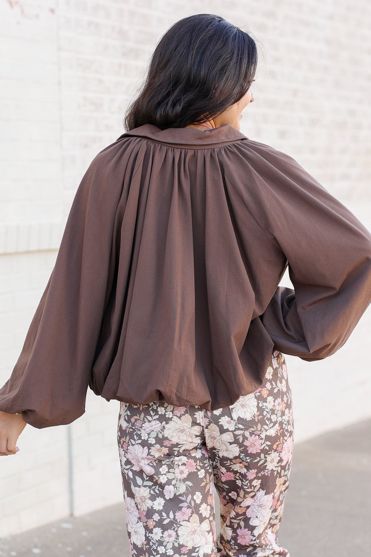 A woman shown from behind wearing a brown long-sleeve blouse with a gathered, balloon-like back and floral pants, standing against a light-colored brick wall.